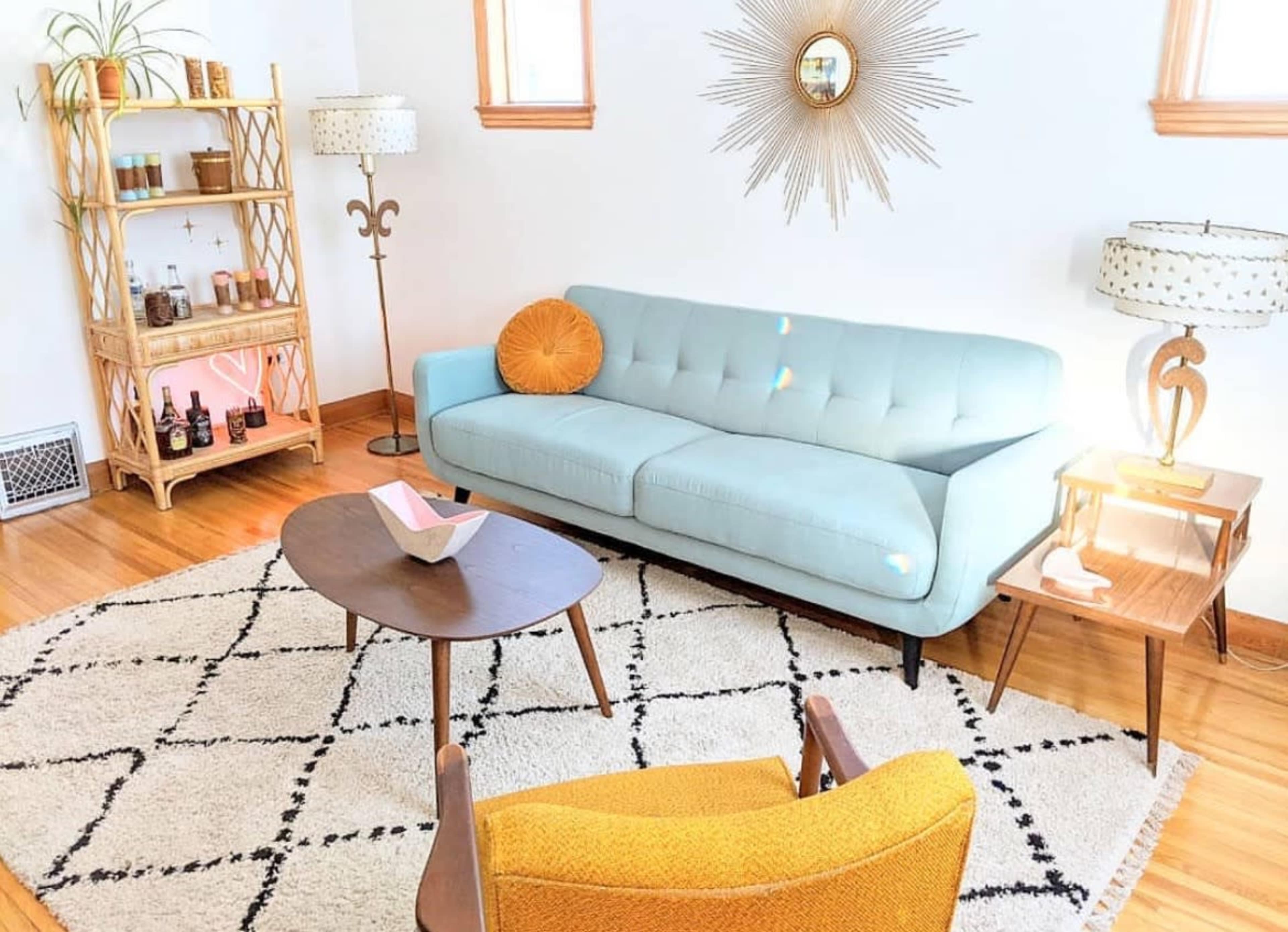 A light-filled living room features a blue tufted sofa, a wooden coffee table, a yellow armchair, and a decorative shelving unit against a white wall.