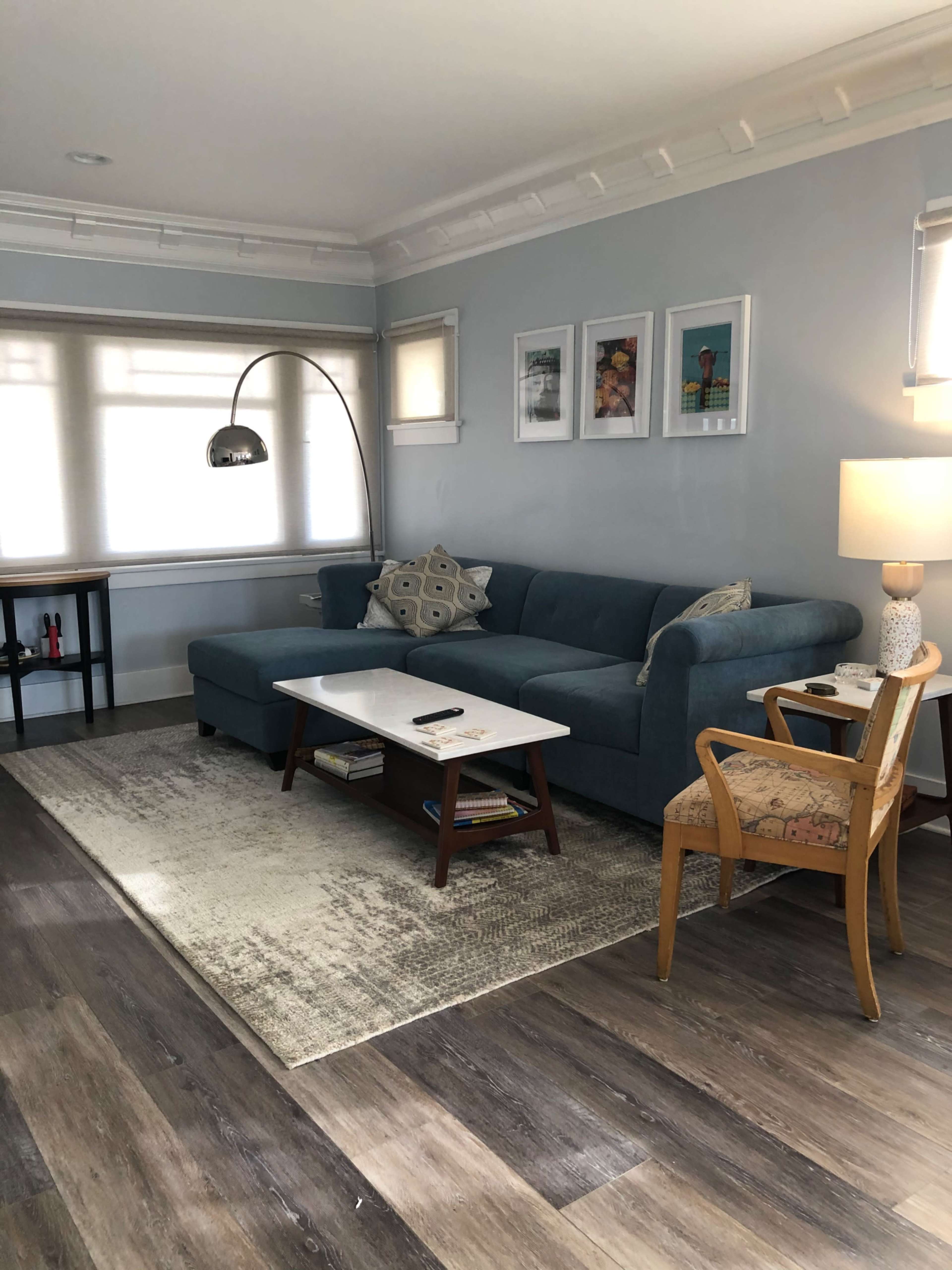 The image shows a modern living room featuring a blue sectional sofa, a marble-top coffee table, and a tall floor lamp, with framed pictures on the wall and a patterned chair in the corner.