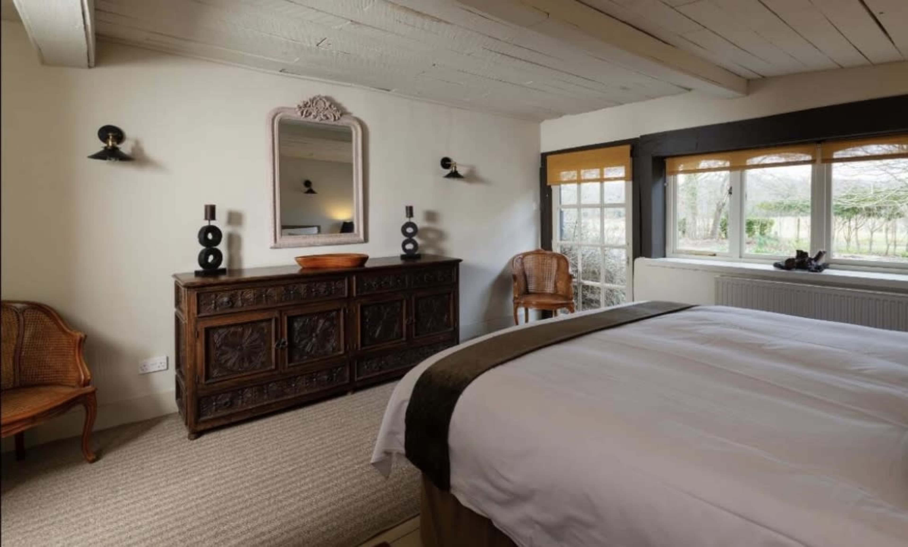 A cozy bedroom featuring a wooden bed, a decorative dresser, two wicker chairs, and a large mirror above the dresser, with a window providing natural light.