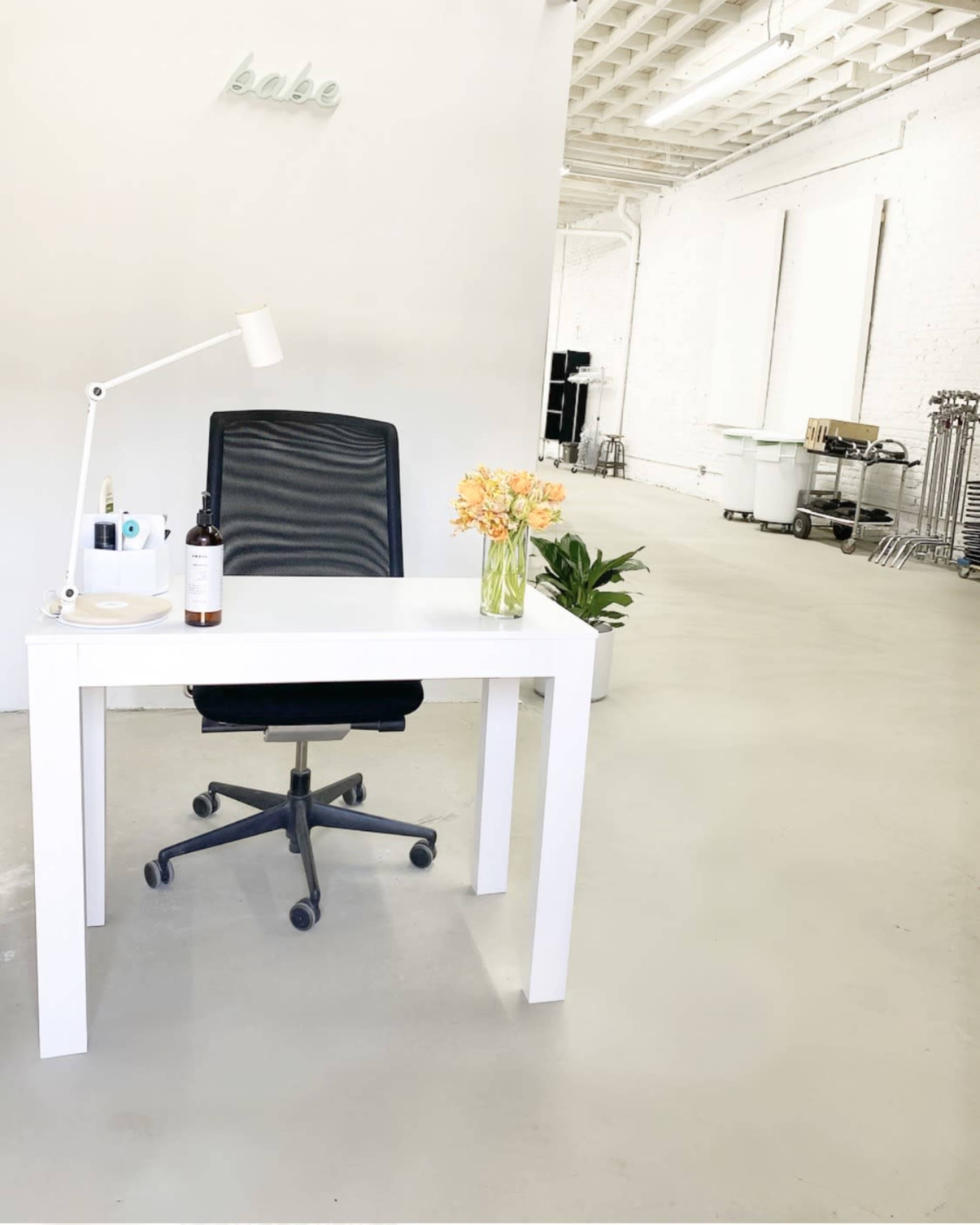 A minimalist office space features a white desk with a black office chair, a vase of flowers, and a lamp, set against a bright, open background.