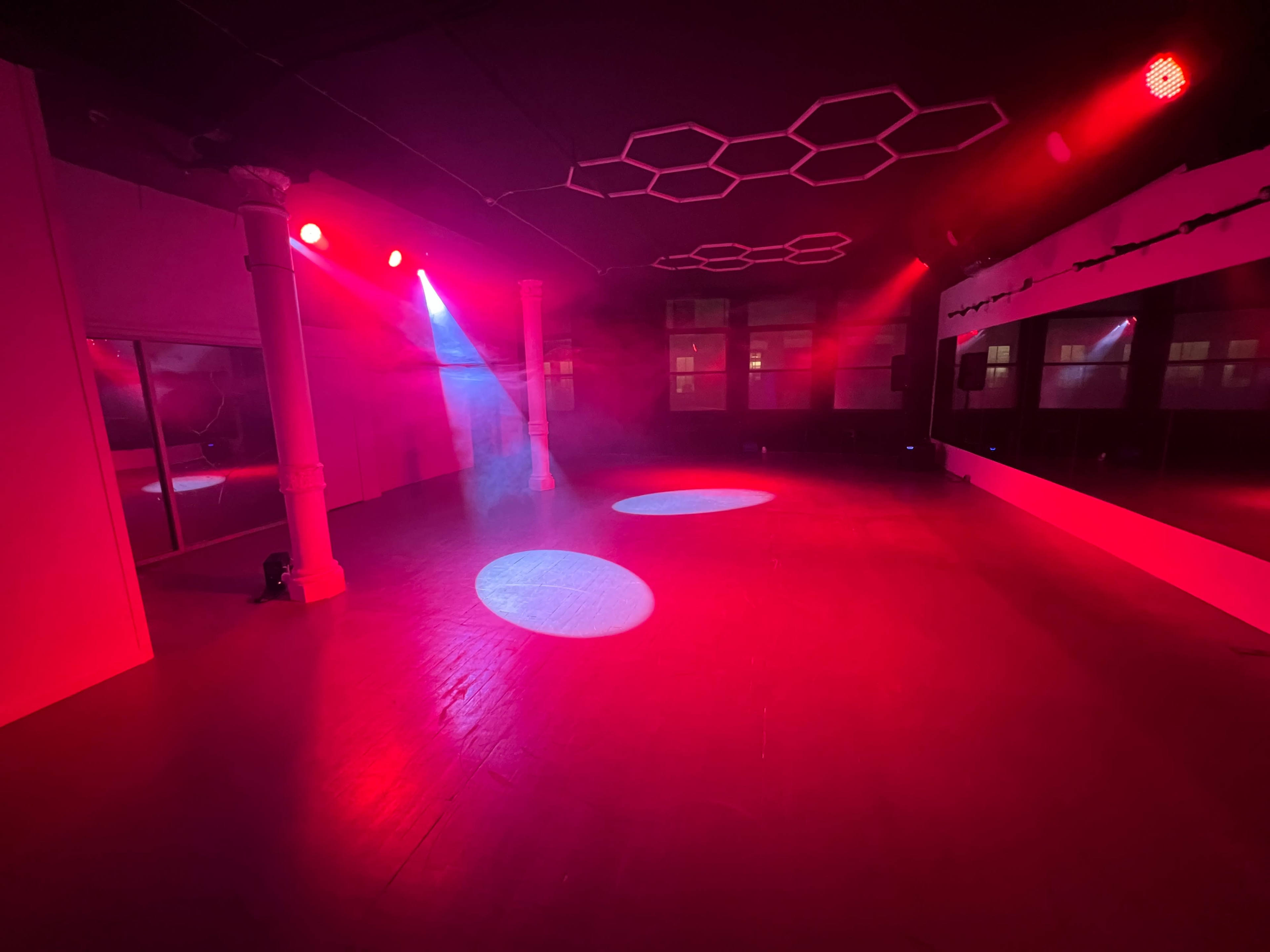 A dance studio is illuminated by red and purple lights, featuring a smooth wooden floor and mirrored walls.