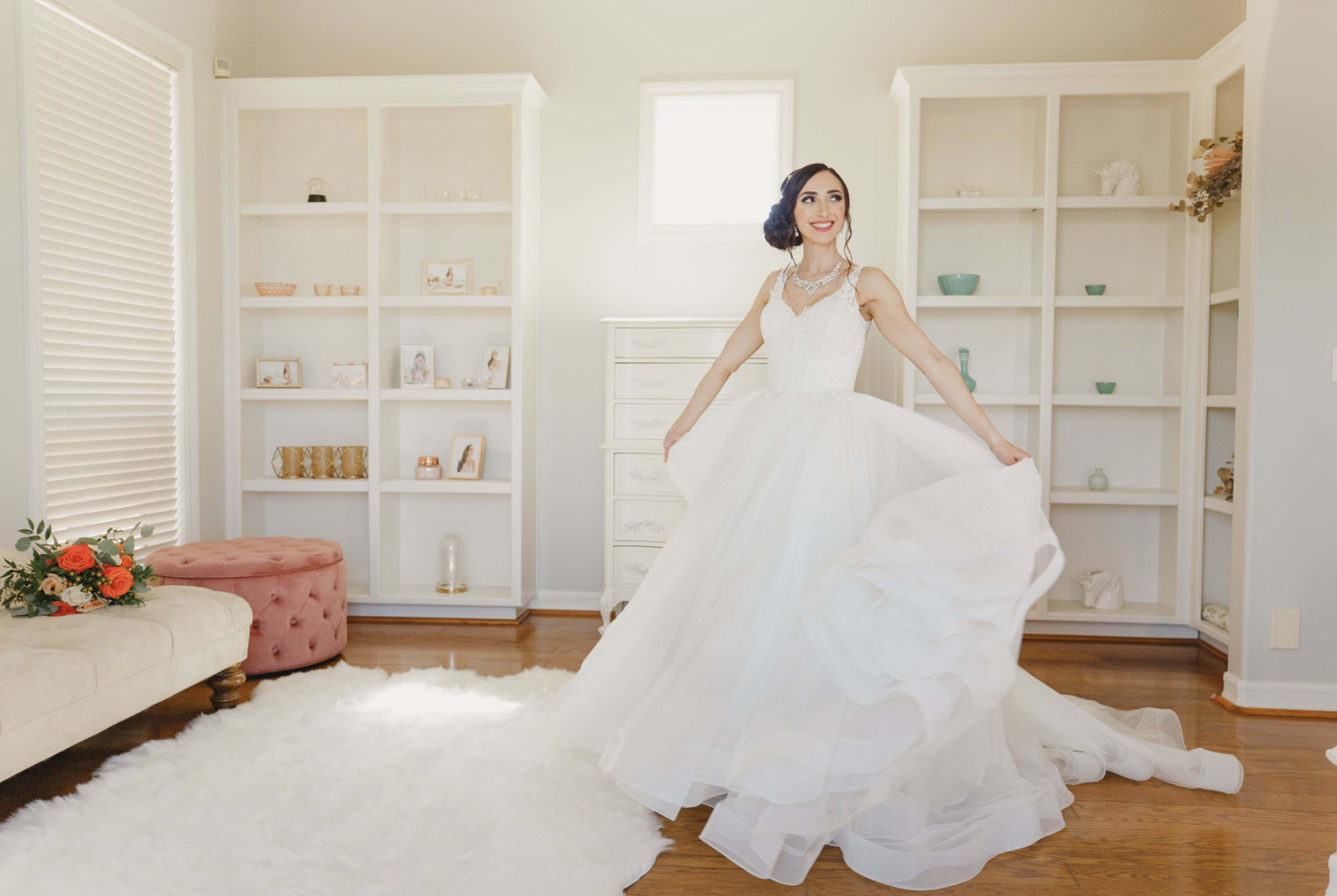 A bride wearing a white gown twirls in a bright room with shelves displaying various decorative items and a plush sofa.