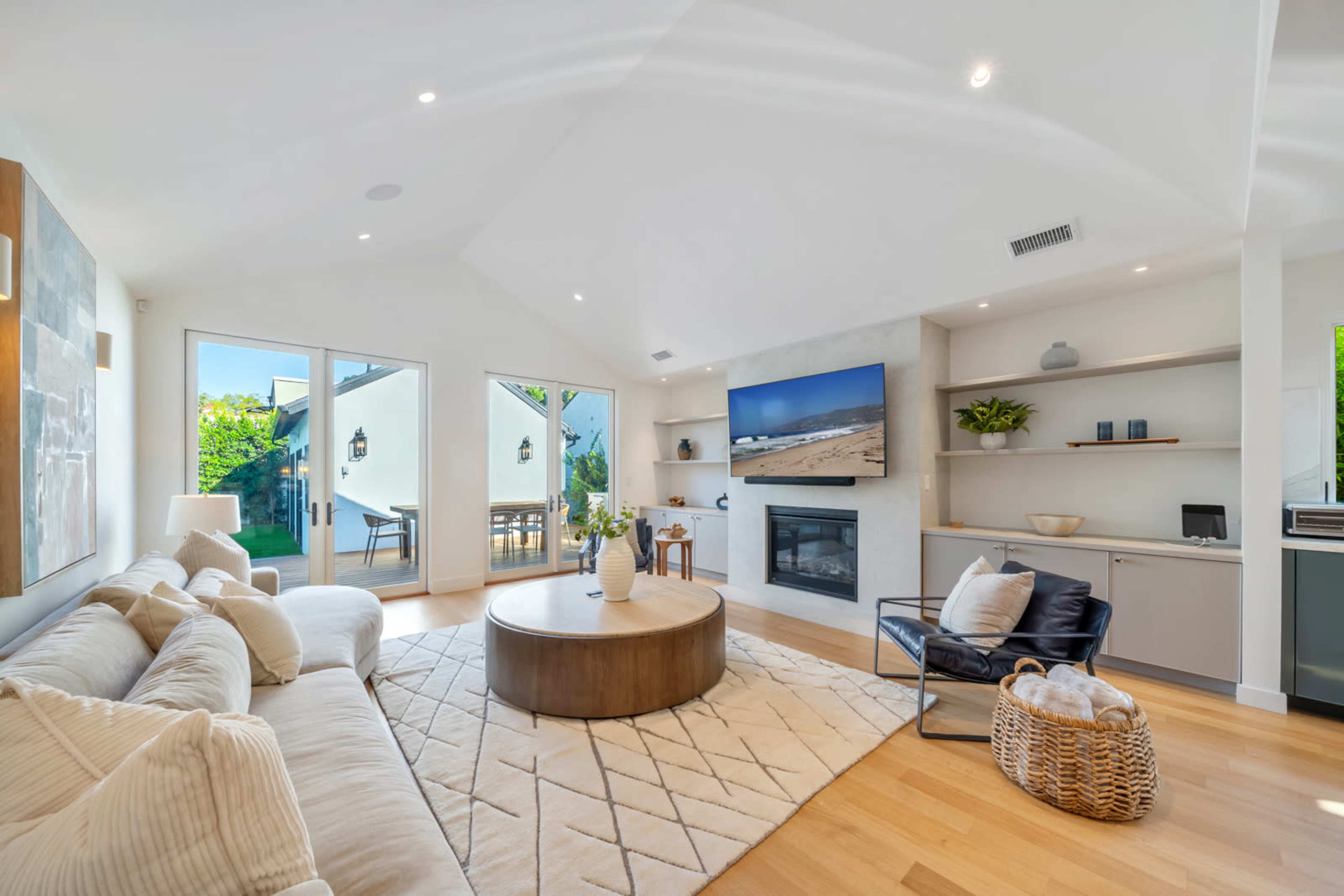 The living room features a light-colored sofa, a circular wooden coffee table, and a television mounted on the wall, with large windows allowing natural light to enter.