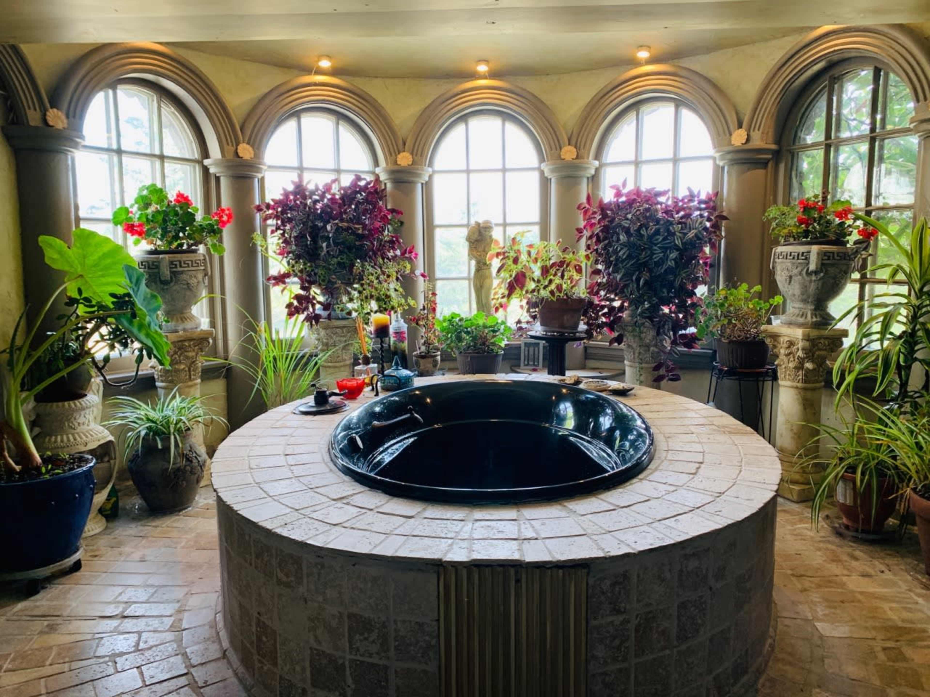 A circular black jacuzzi is surrounded by ornate windows adorned with potted plants and flowers in a well-lit room.