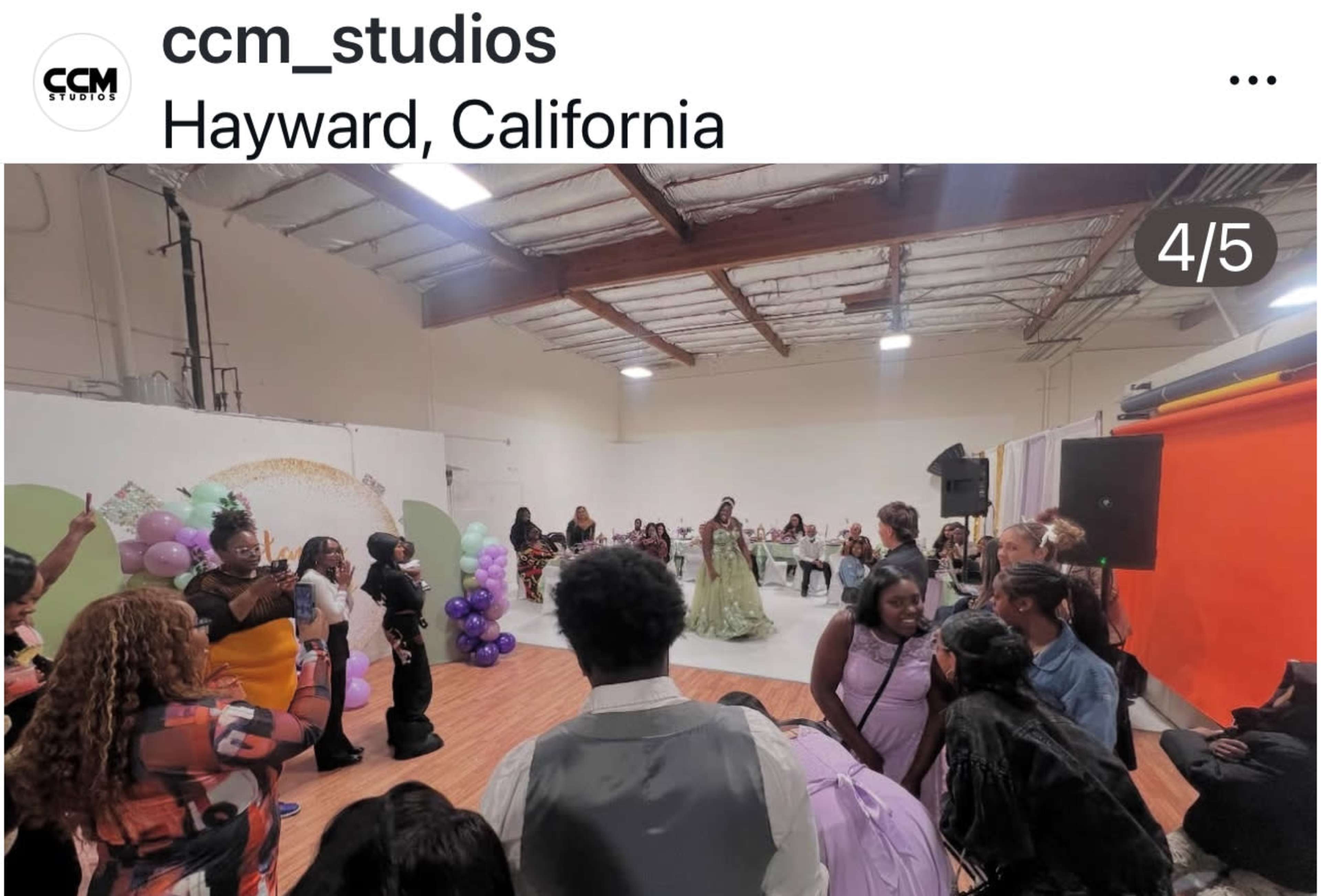 The image shows a studio event in Hayward, California, where models walk down a runway surrounded by an audience in a decorated space.