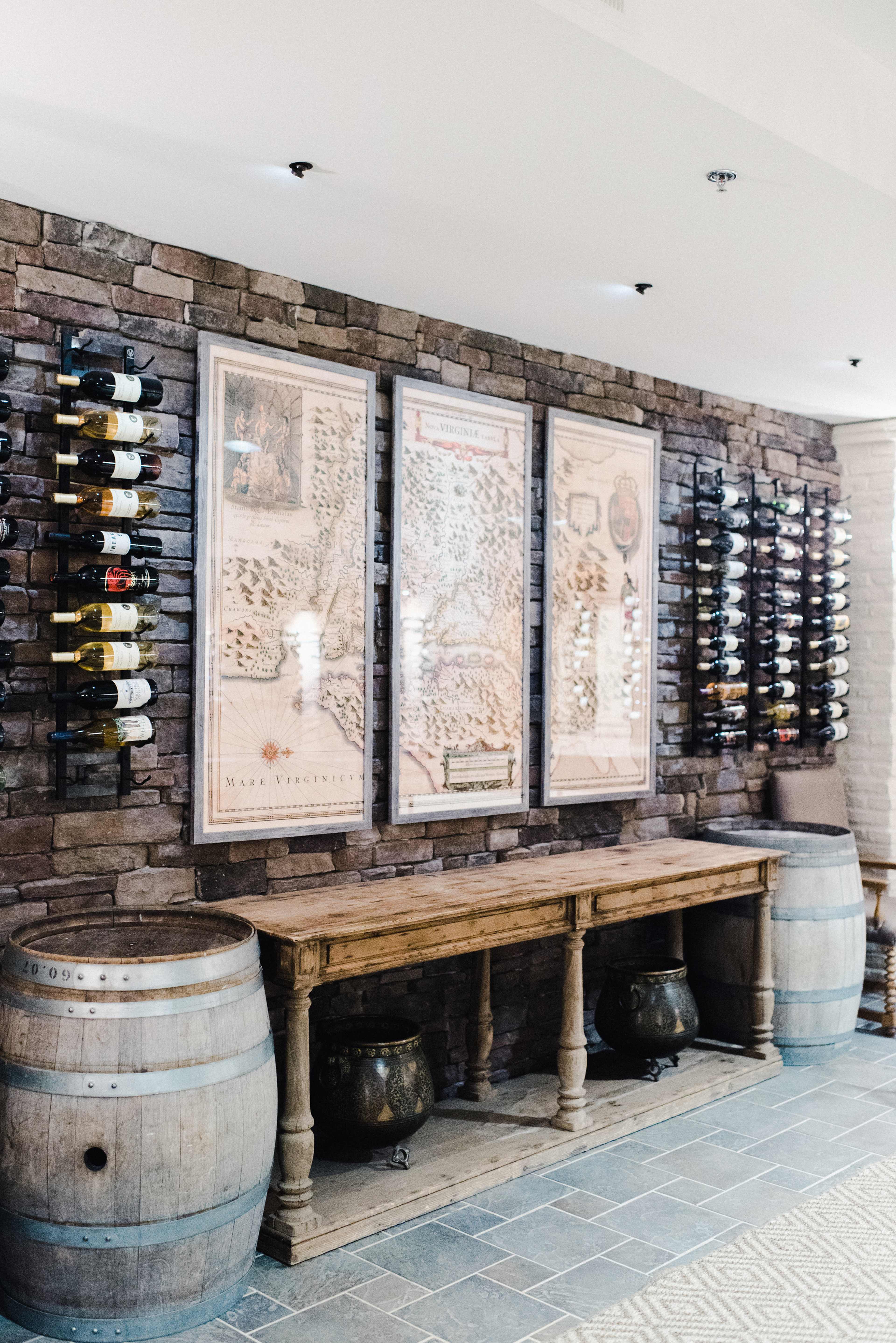 A wine cellar with a stone wall, wooden table, two wine barrels, and several bottles of wine displayed on racks and framed maps on the wall.