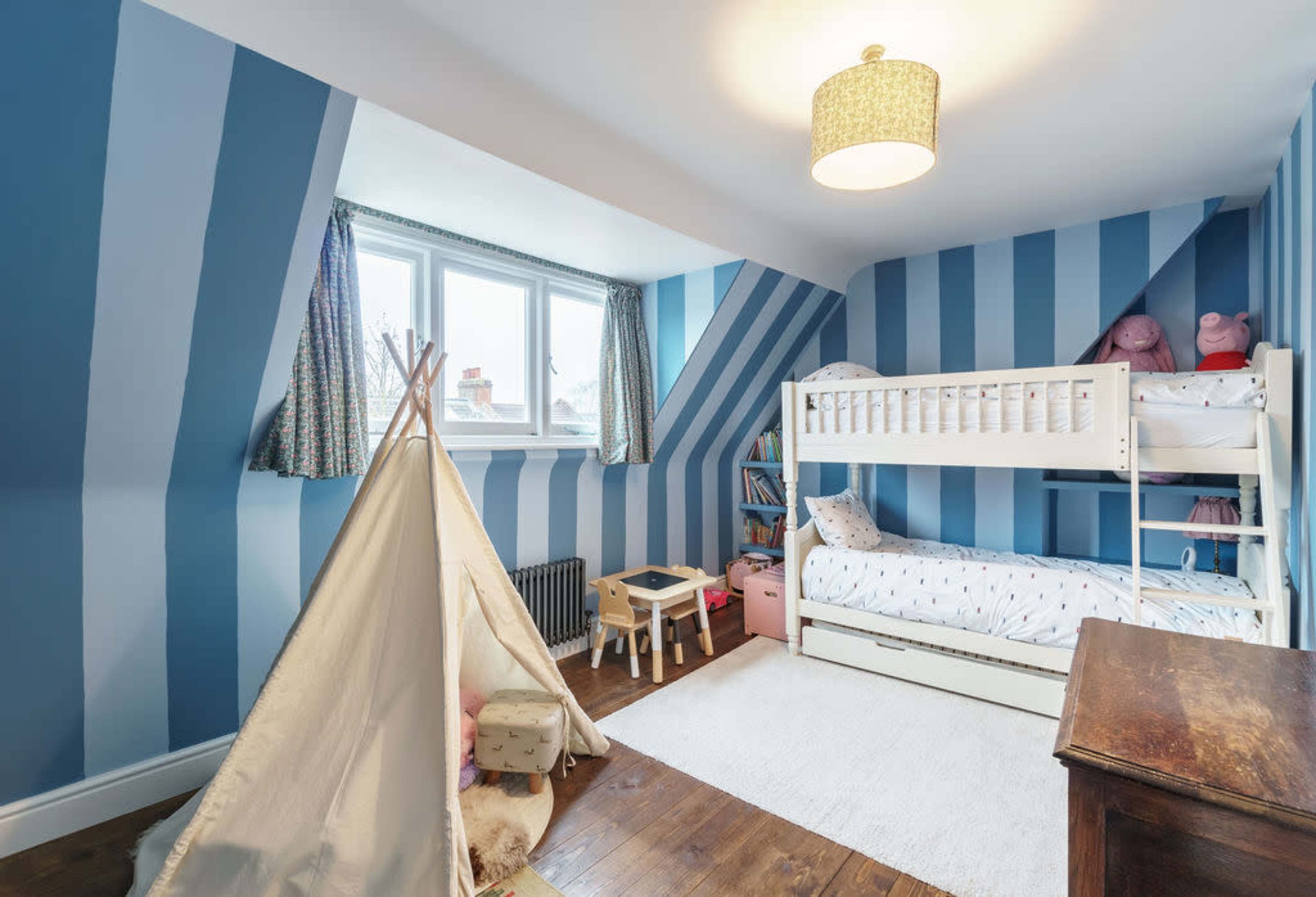 A children's bedroom features striped blue walls, a bunk bed, a play tent, and a cozy rug on a wooden floor.