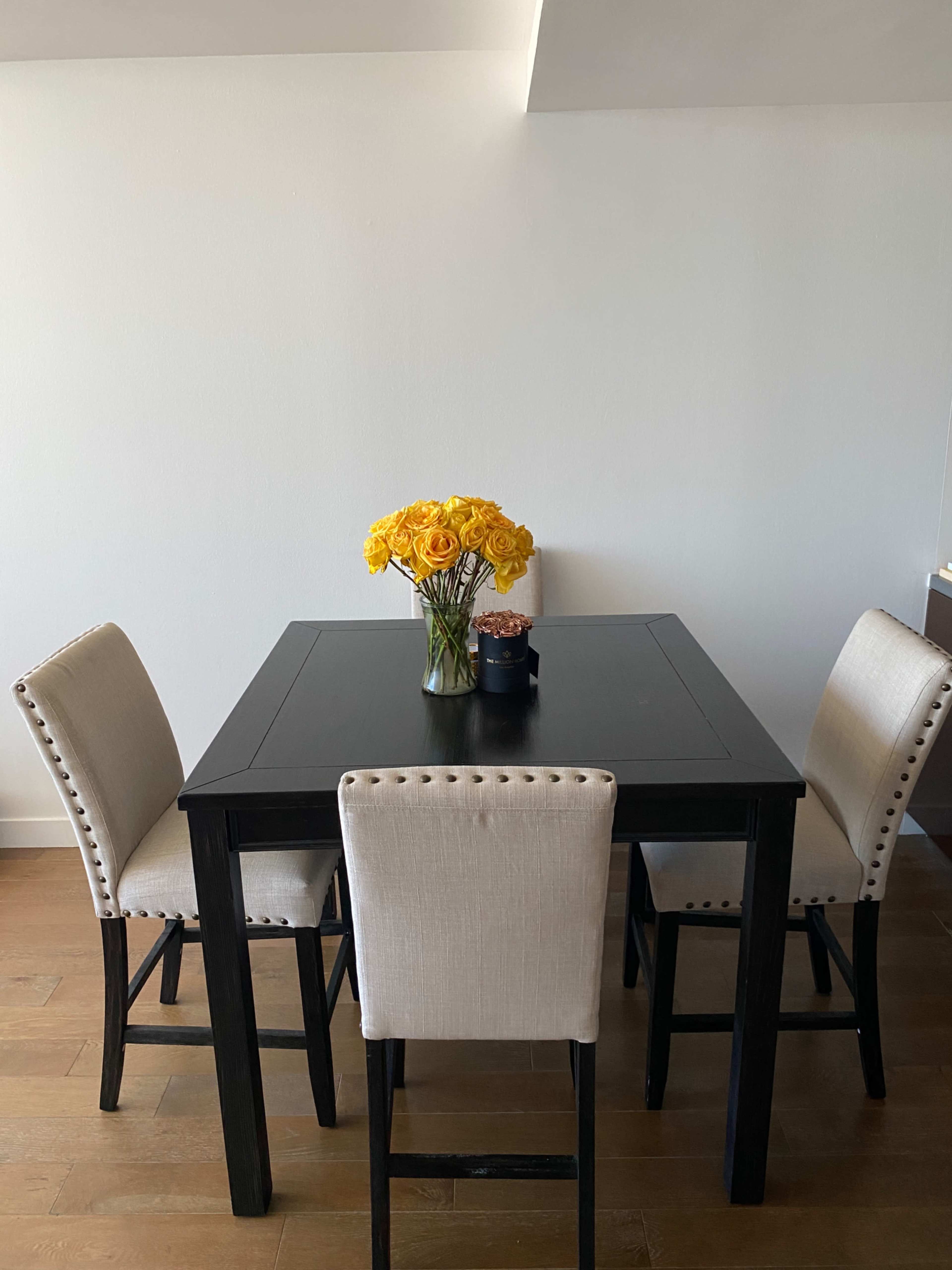 A square black table is set with four beige upholstered chairs, and a vase of yellow roses is placed in the center.