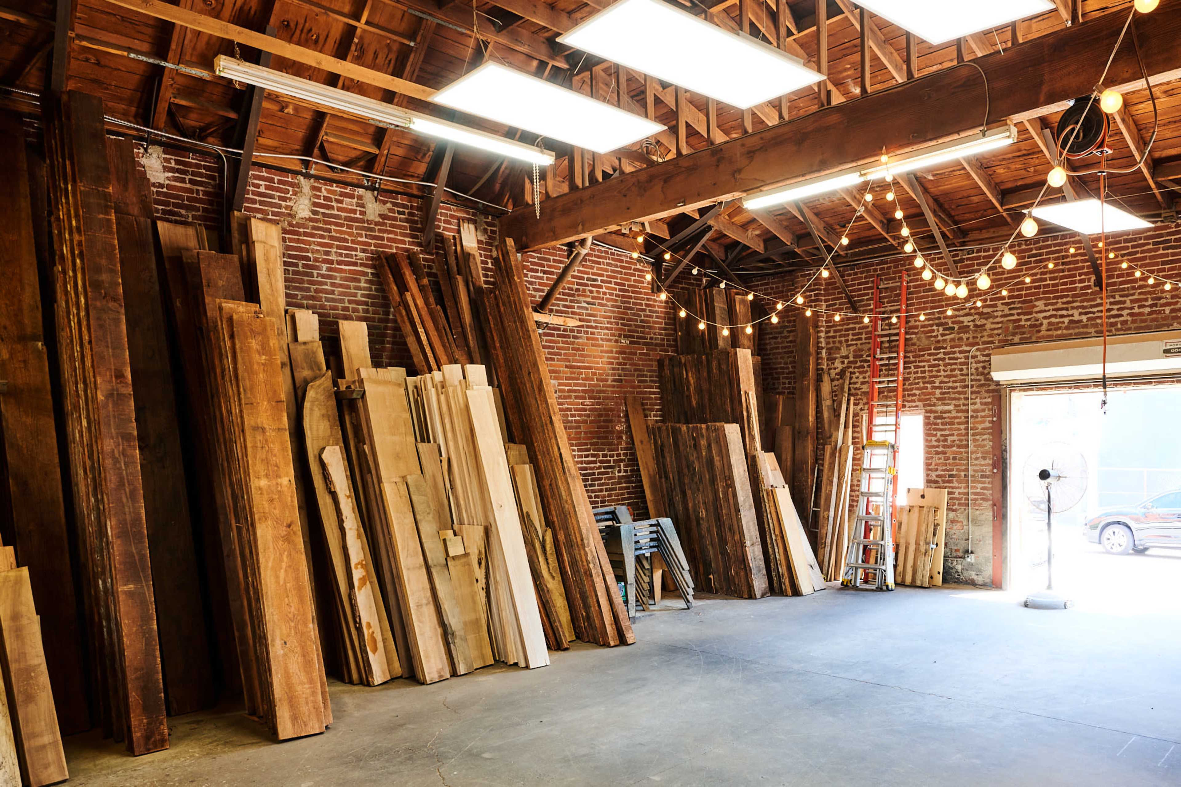 A spacious workshop with stacked wooden planks against the brick walls and overhead string lights illuminating the area.
