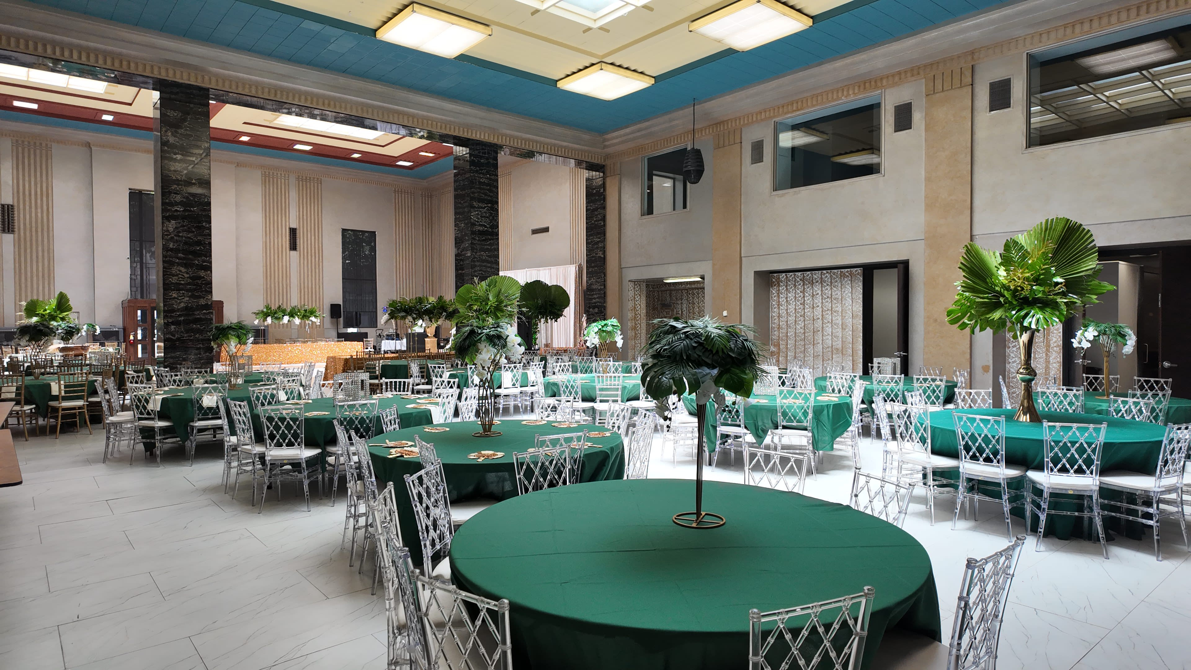 The image shows a spacious indoor venue set up for an event, featuring round tables covered with green tablecloths and surrounded by silver chairs, alongside large potted plants.