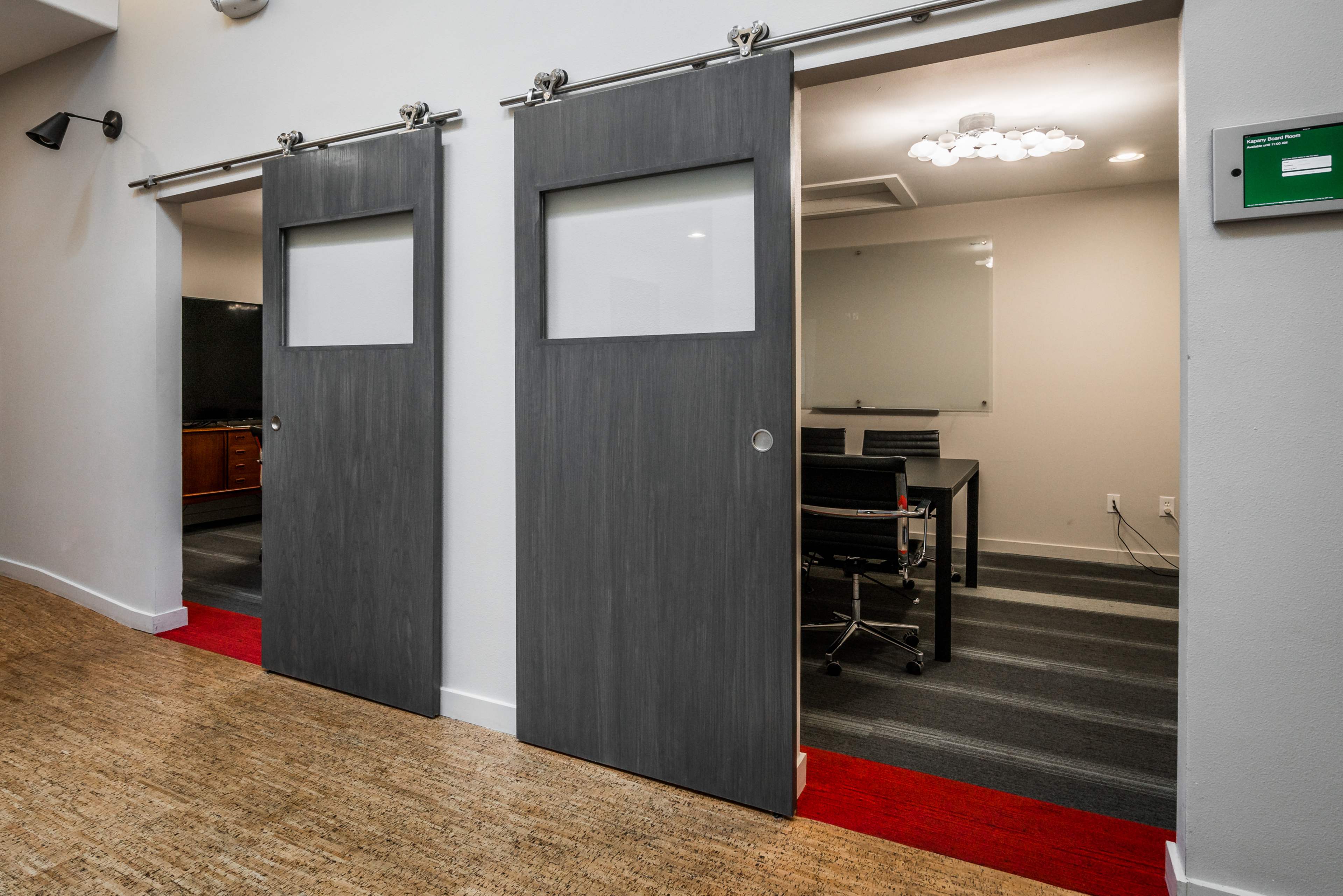 Two sliding barn doors with glass panels lead to a meeting room and an office in a modern interior space.