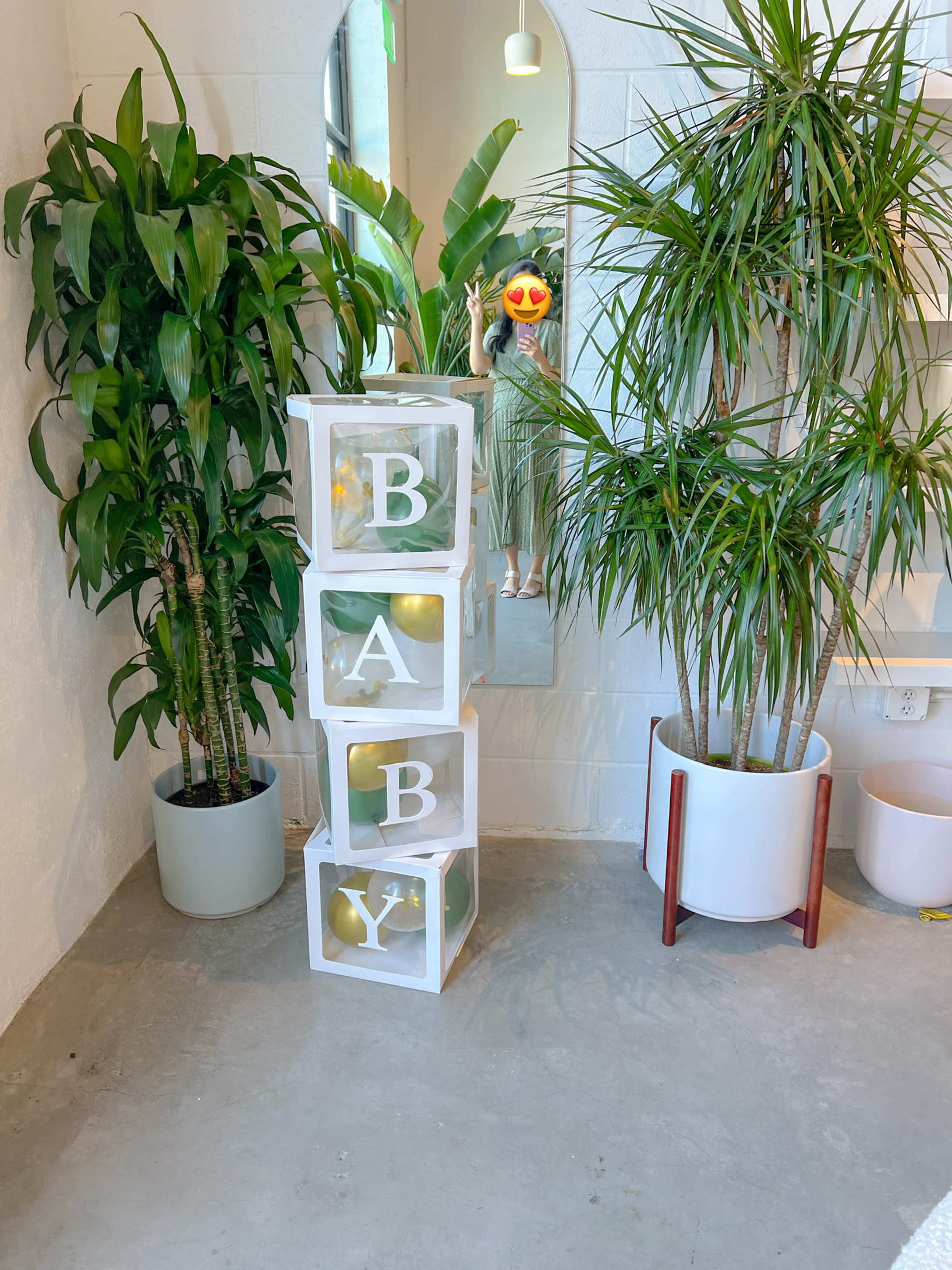 A stack of clear boxes with the letters "B A B Y" arranged in a bright, modern space featuring potted plants and a large mirror.
