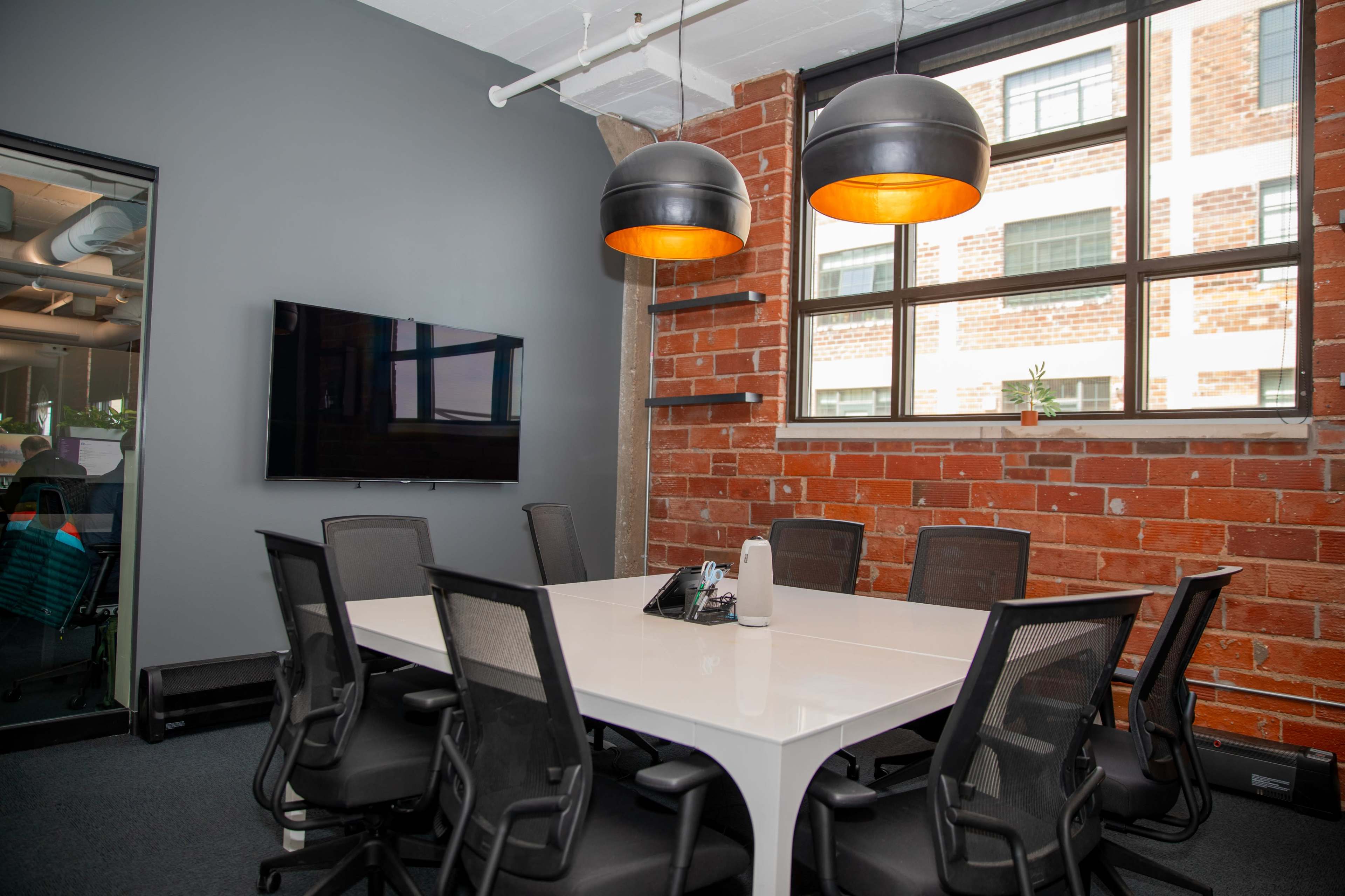 The image shows a modern conference room with a large white table, black mesh chairs, and two pendant lights, all set against a backdrop of exposed brick walls.