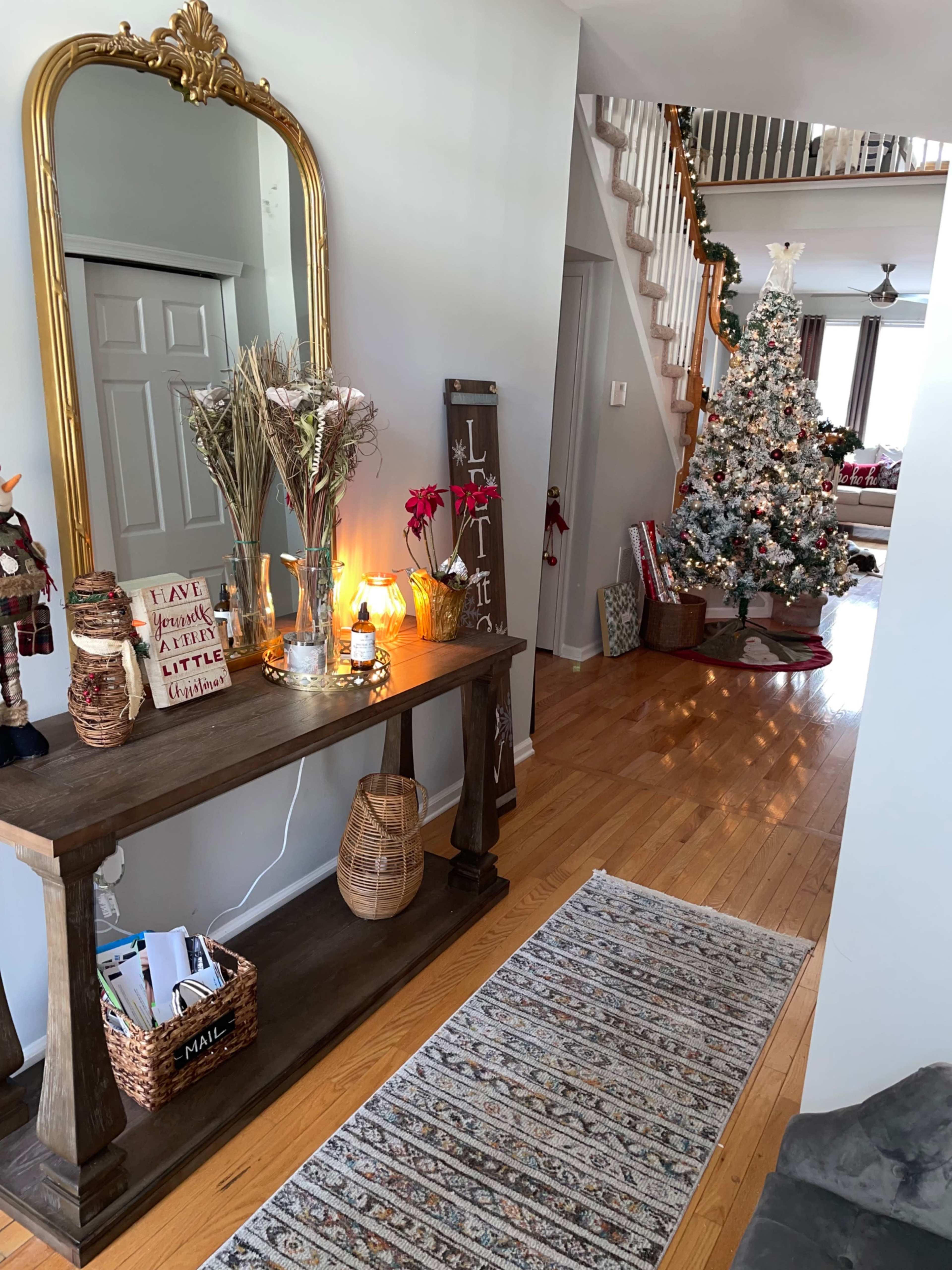 A hallway with a decorated Christmas tree in the background, a console table with decor and a mirror, and a staircase leading to the upper floor.