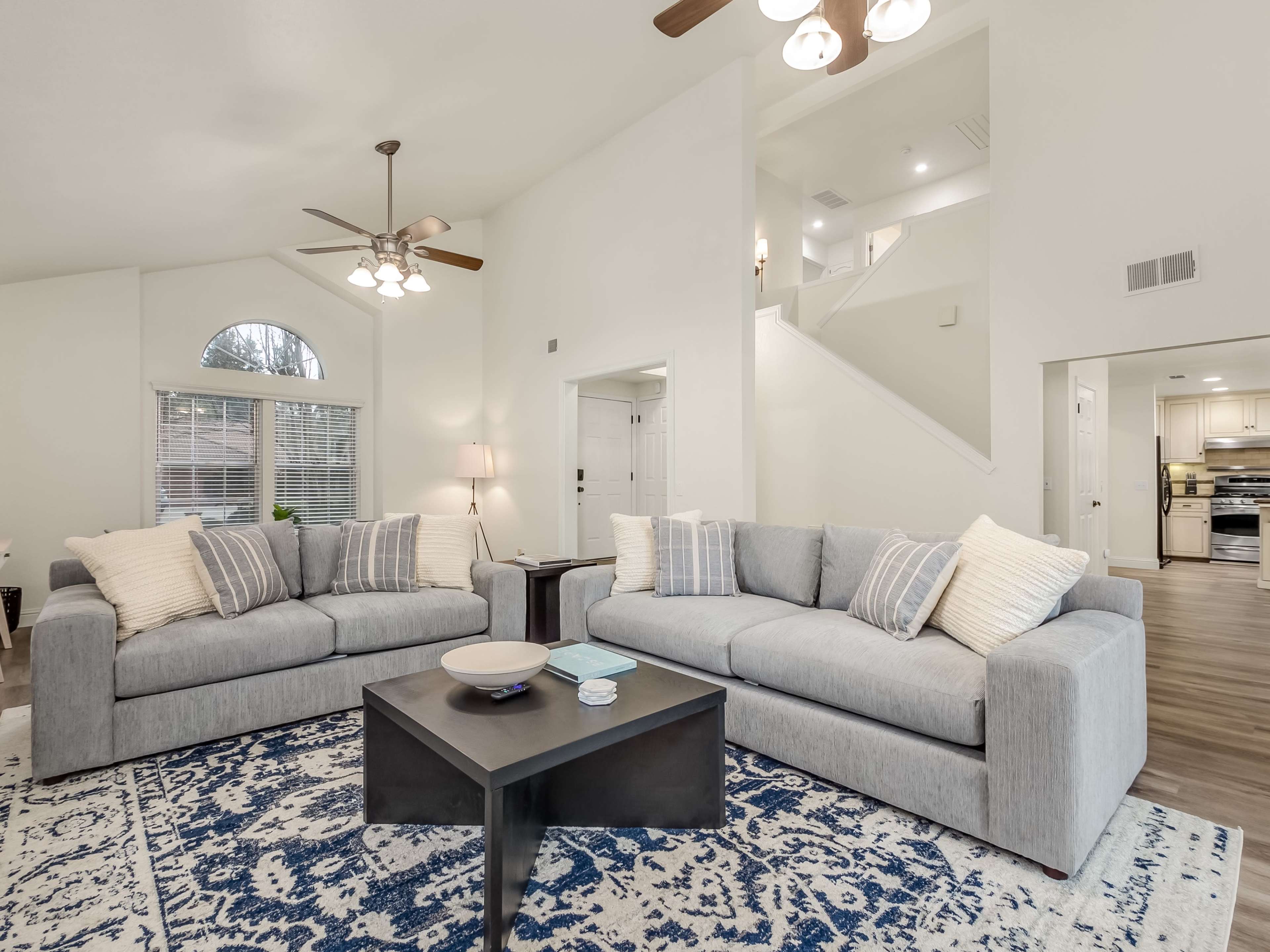 The living room features two gray sofas facing a dark coffee table, situated on a patterned area rug, with light streaming in through large windows.