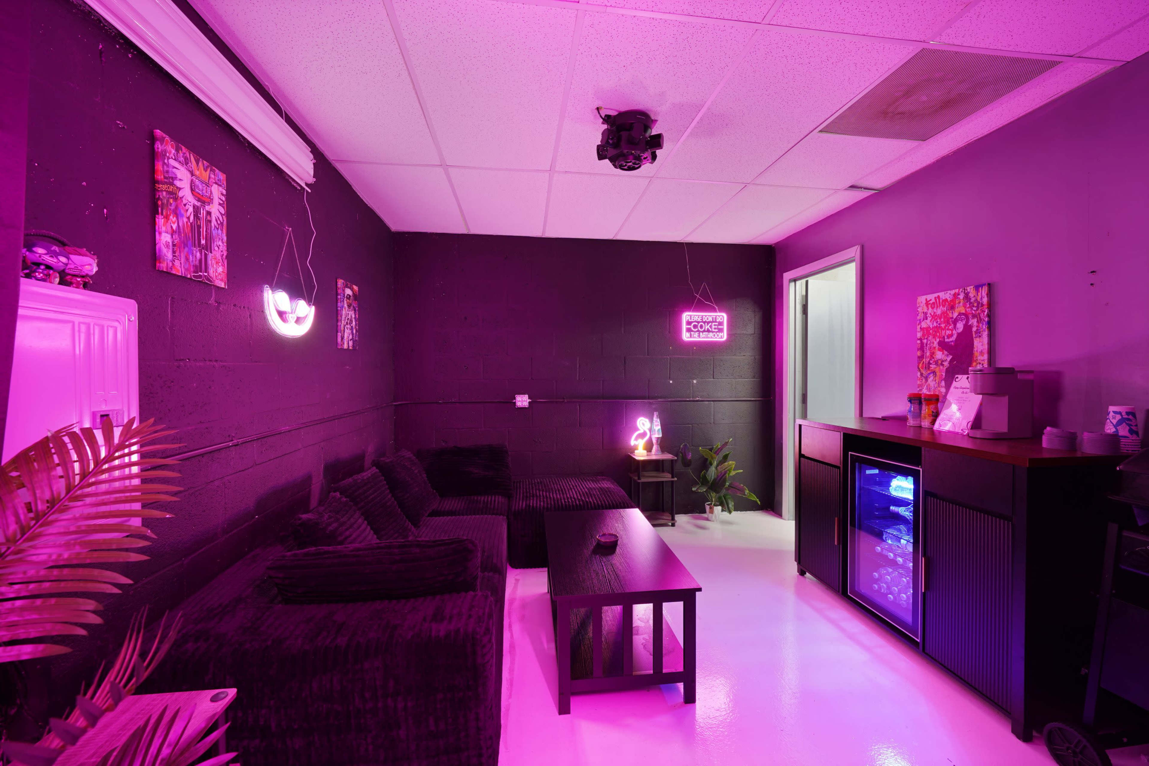 The image shows a dimly lit lounge area with purple walls, a black sofa, a coffee table, a mini fridge, and neon signs.