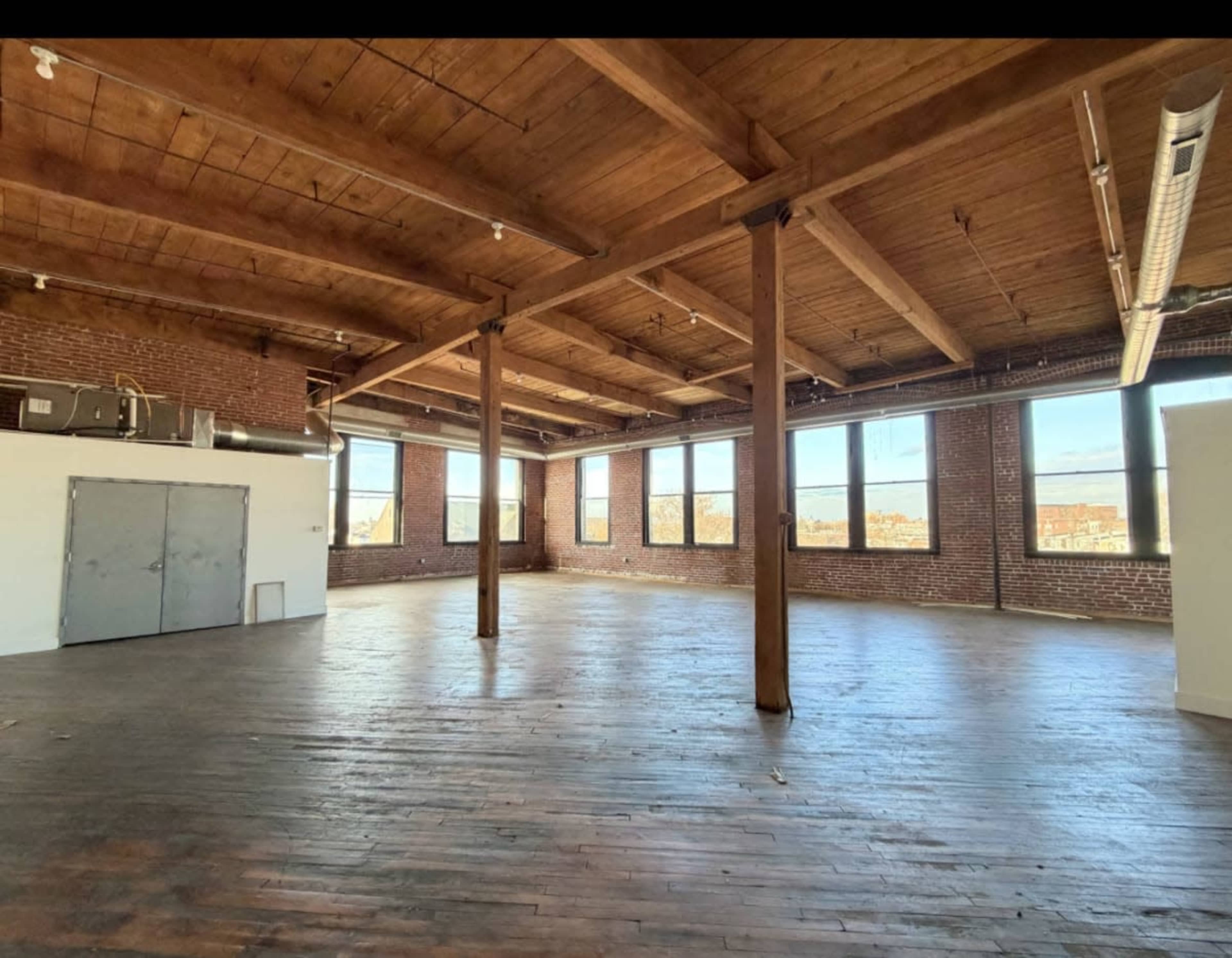 The image shows a spacious loft with wooden beams, large windows, and a polished wooden floor.