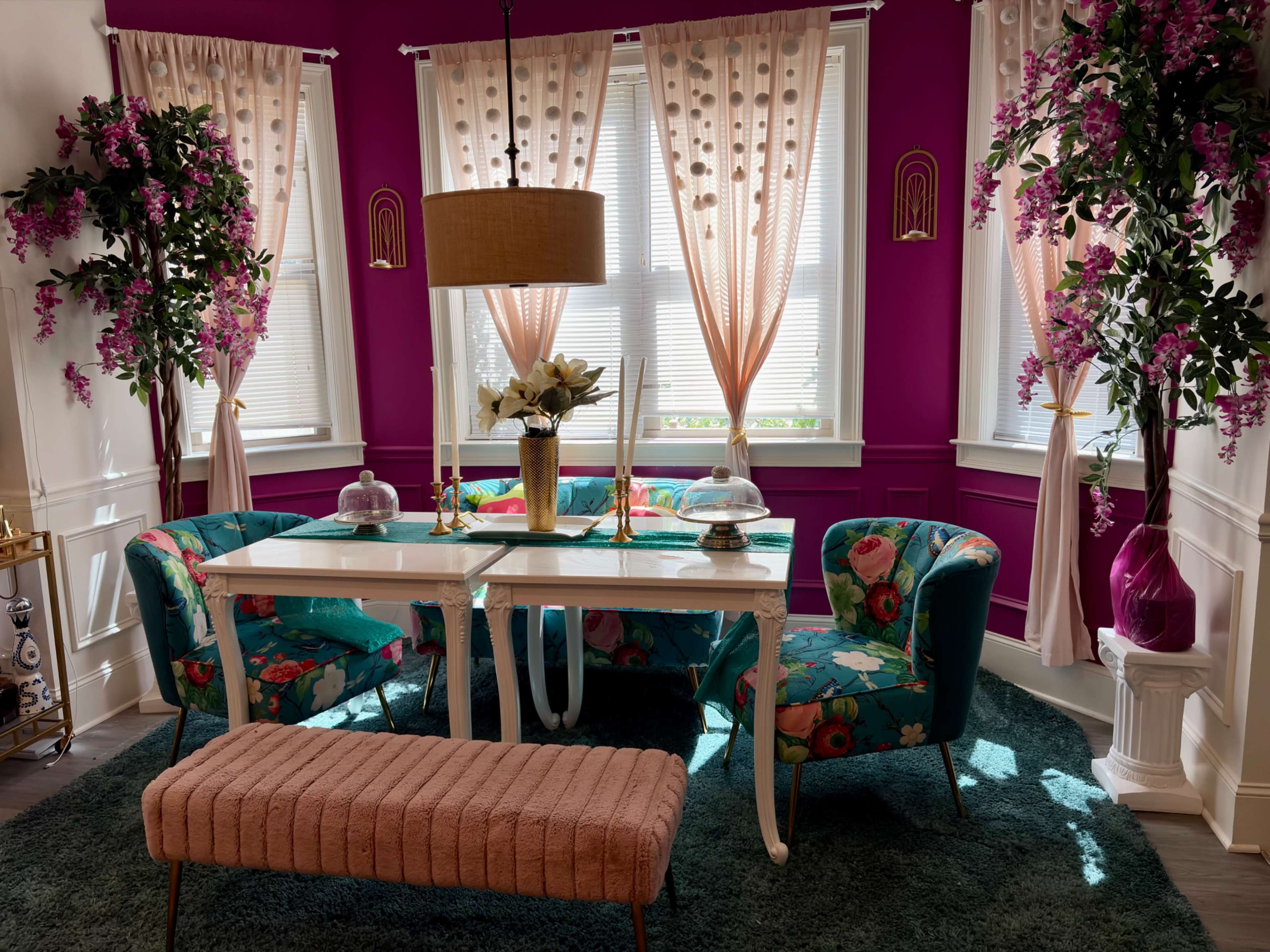 The image shows a brightly colored dining nook with floral patterned seating, a white table, and decorative accents, set against purple walls and large windows.
