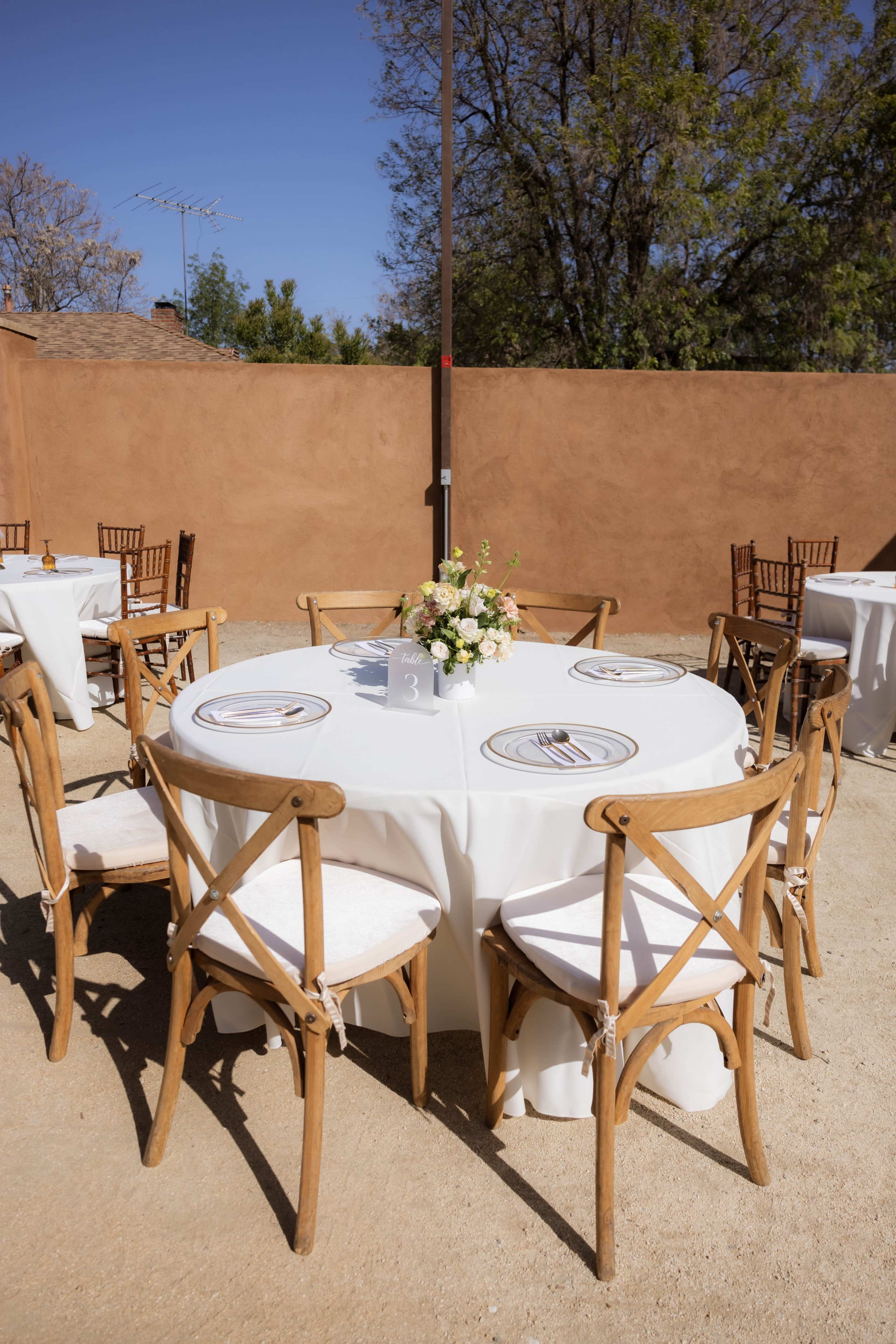 A round table with white tablecloth and floral centerpiece is set for an outdoor event, surrounded by wooden chairs against a backdrop of a beige wall and blue sky.