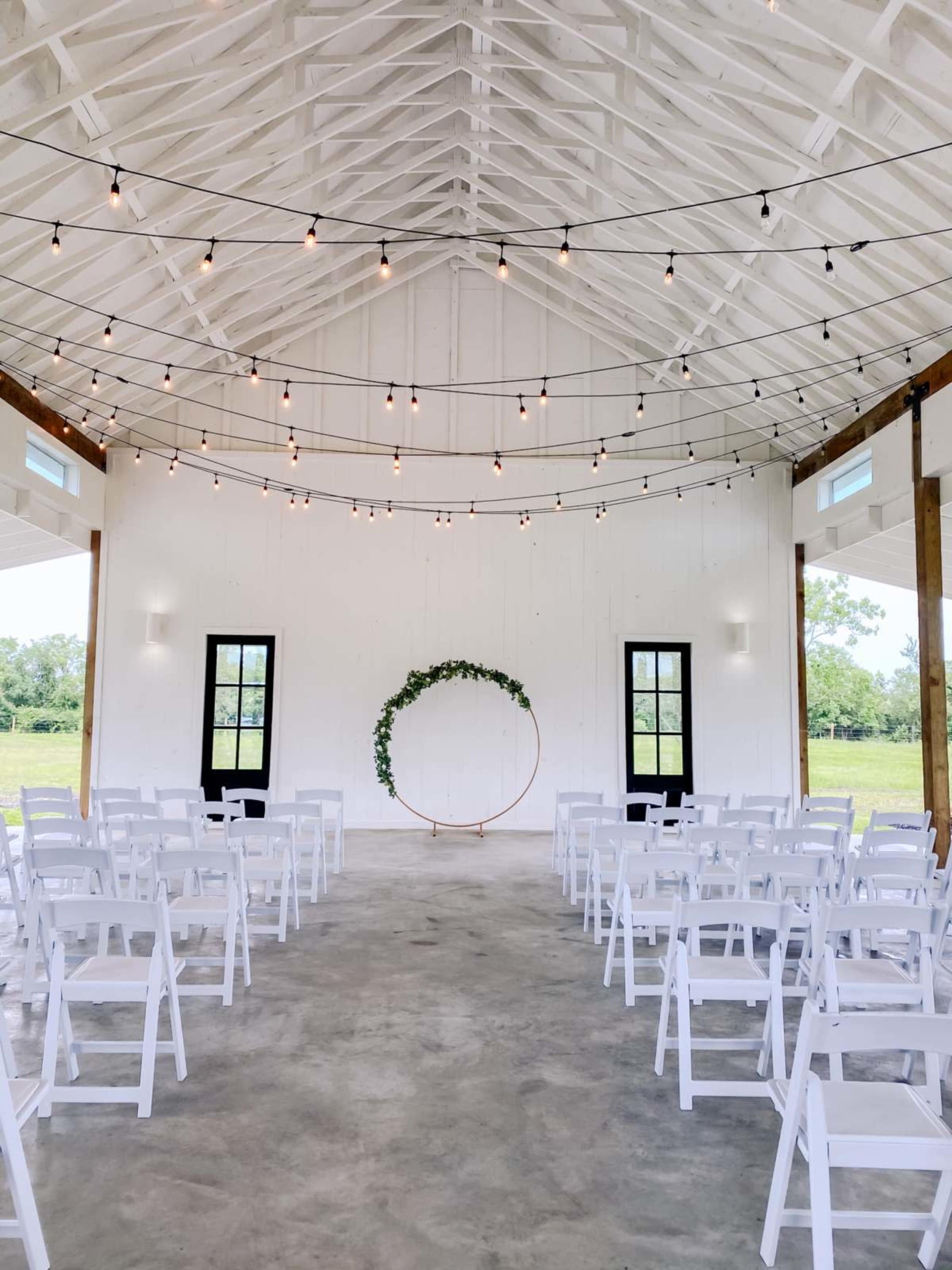 The image shows a wedding venue with rows of white chairs facing a circular floral arch under a string light canopy in a white-walled building.