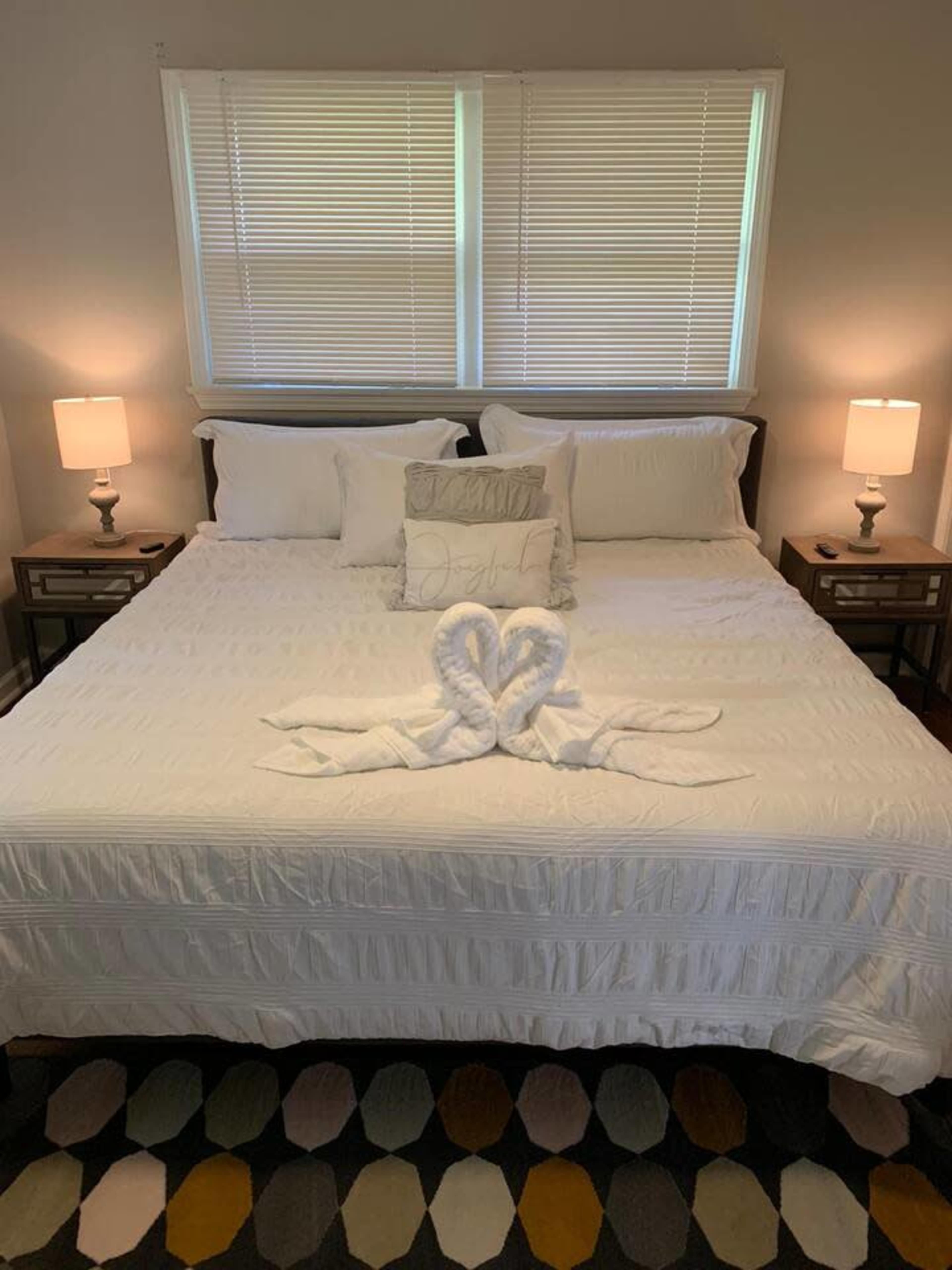 A neatly made bed with decorative pillows and towel sculptures sits between two bedside tables and lamps in a well-lit room.