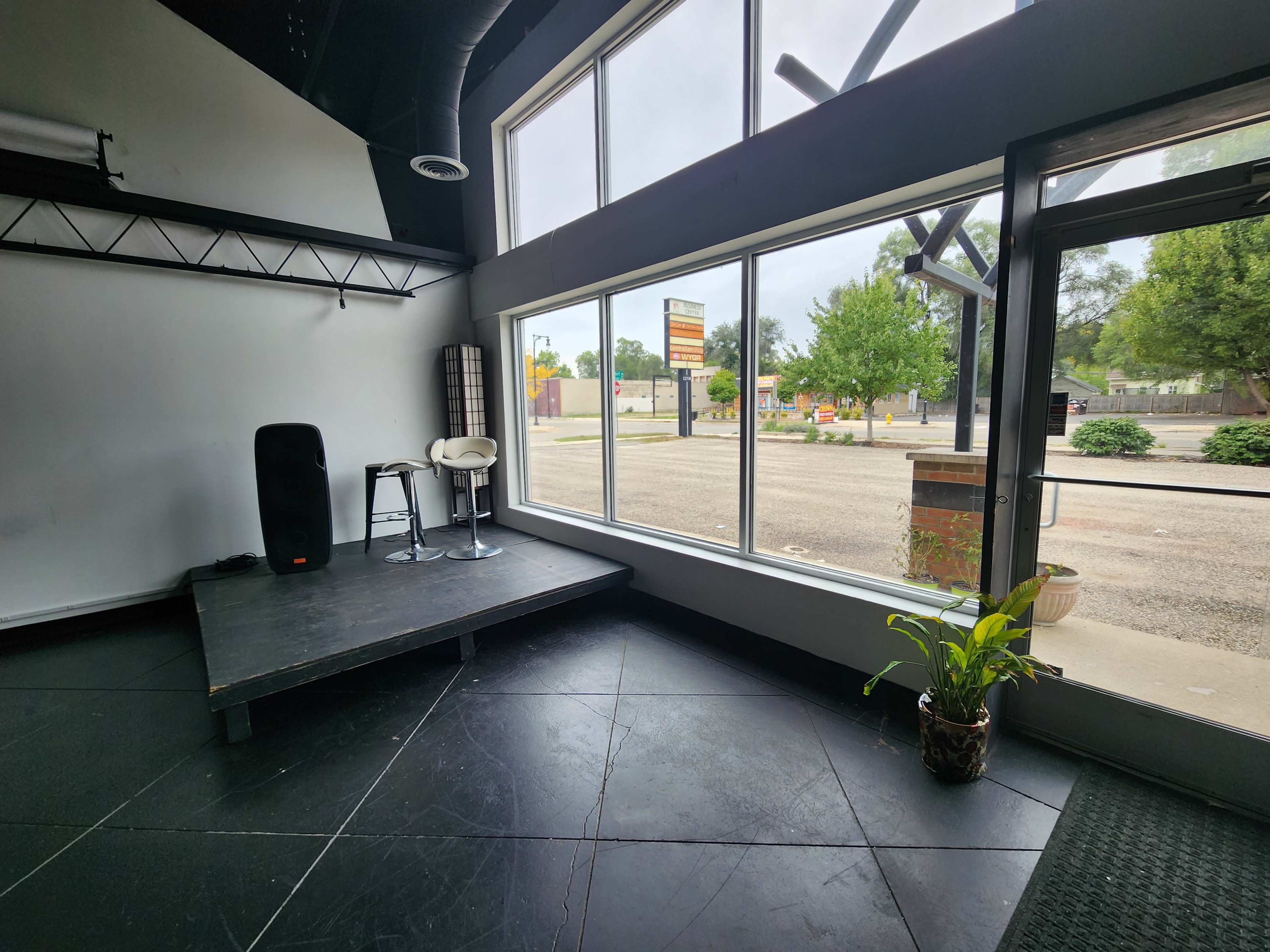 An empty performance space with a small stage, two stools, and large windows facing an outdoor area with a parking lot and greenery.