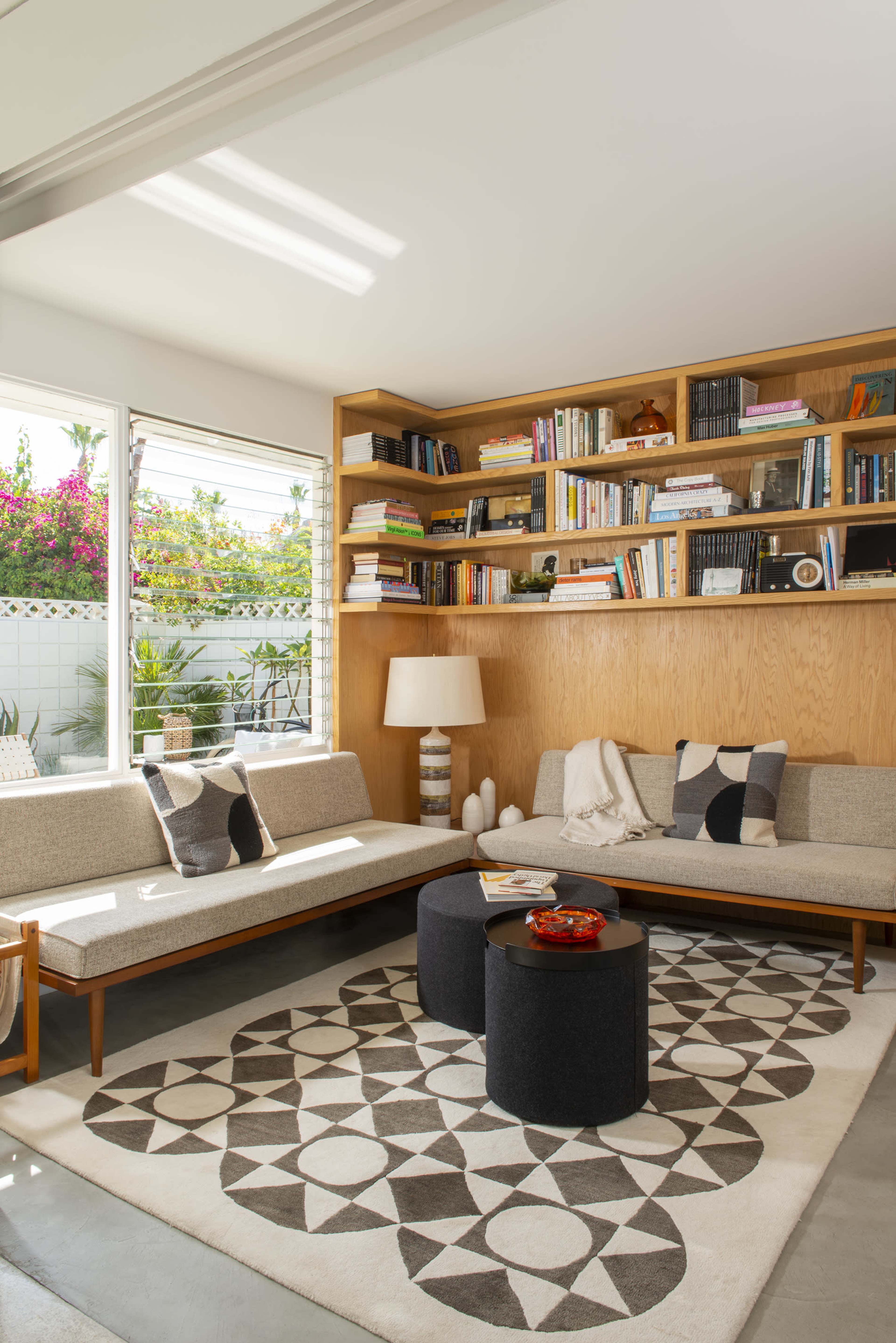 A modern, minimalistic living room featuring a wooden bookshelf filled with books, a cozy seating area, and a circular coffee table on a patterned rug.
