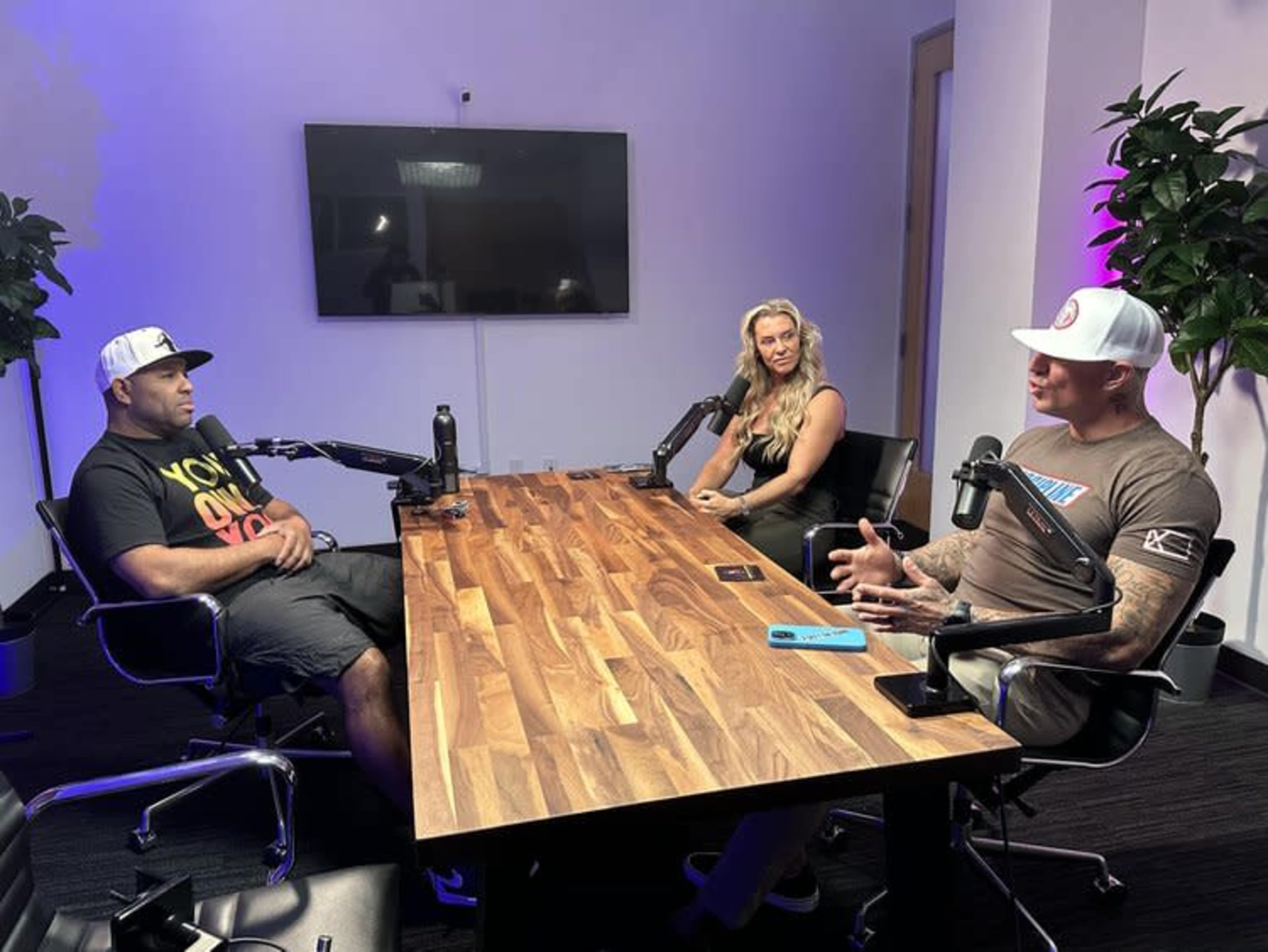 Three people are seated at a wooden table in a modern meeting room with a large screen in the background.