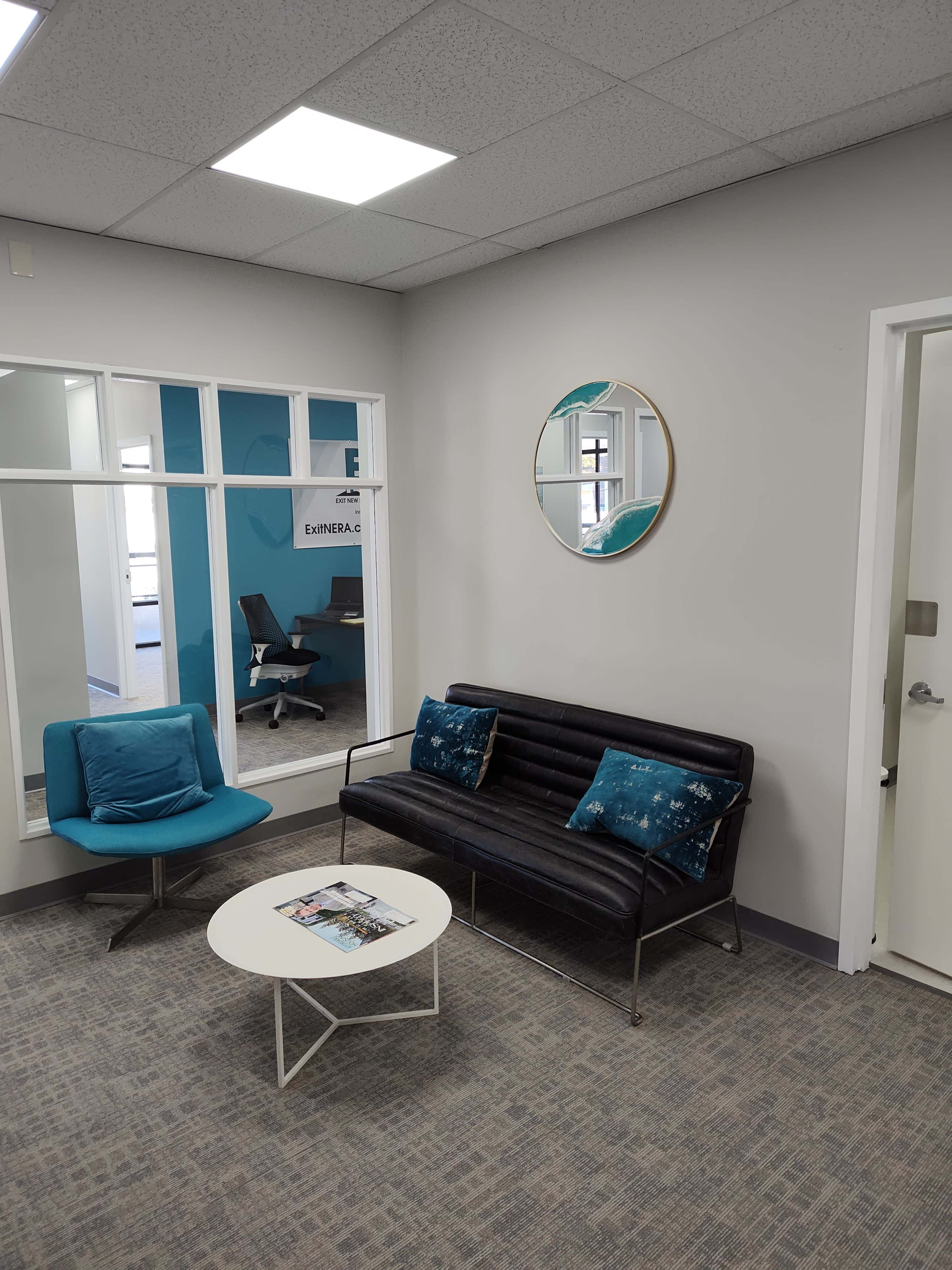 The image shows a waiting area with a black couch and a light blue chair, accompanied by a white coffee table, in a well-lit office space.