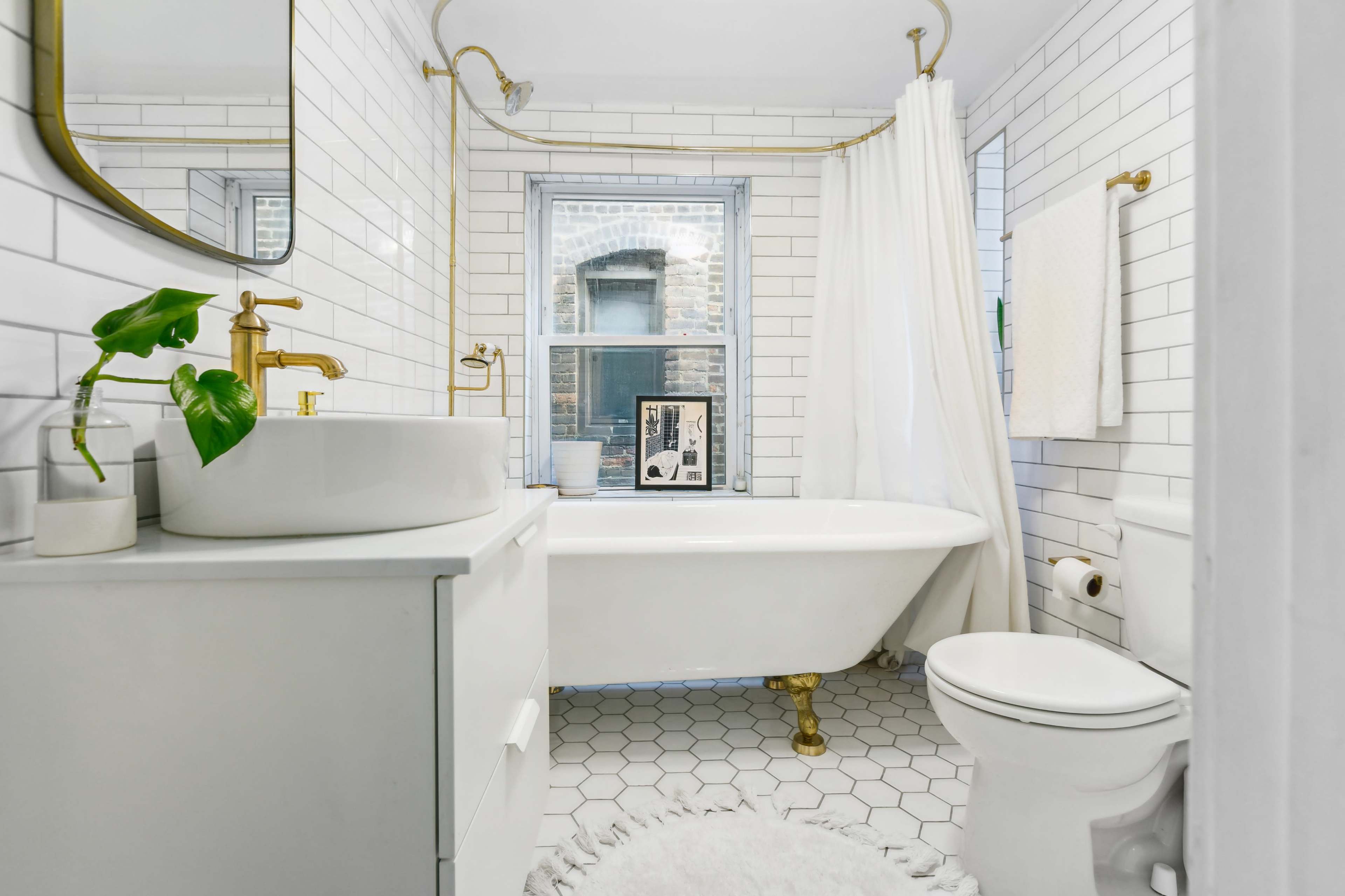 A modern bathroom features a freestanding tub, white tiled walls, a gold-accented mirror, and a large window allowing natural light.