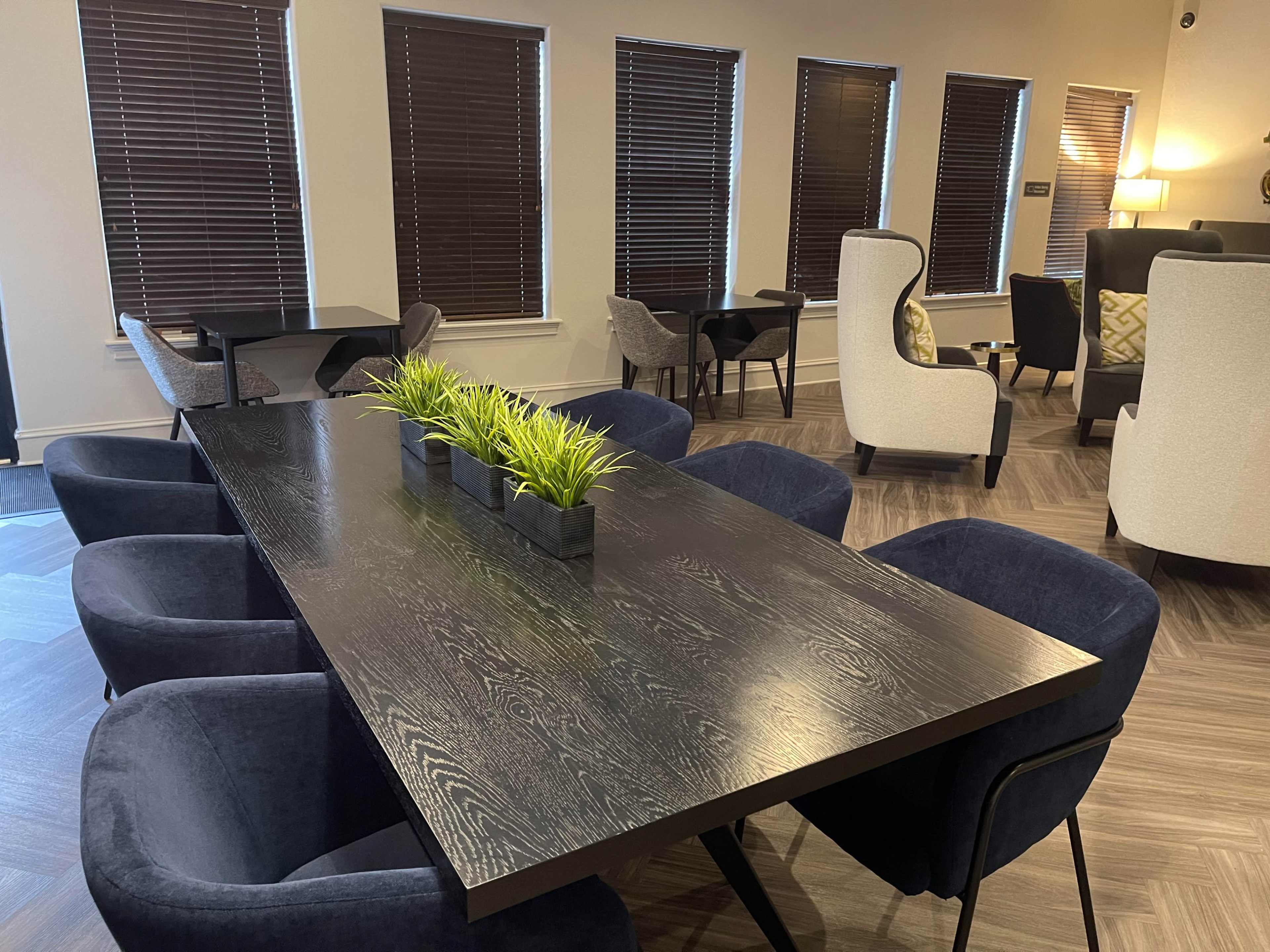 A large dark wooden table sits in a modern room surrounded by blue upholstered chairs and potted plants, with windows covered by dark blinds.