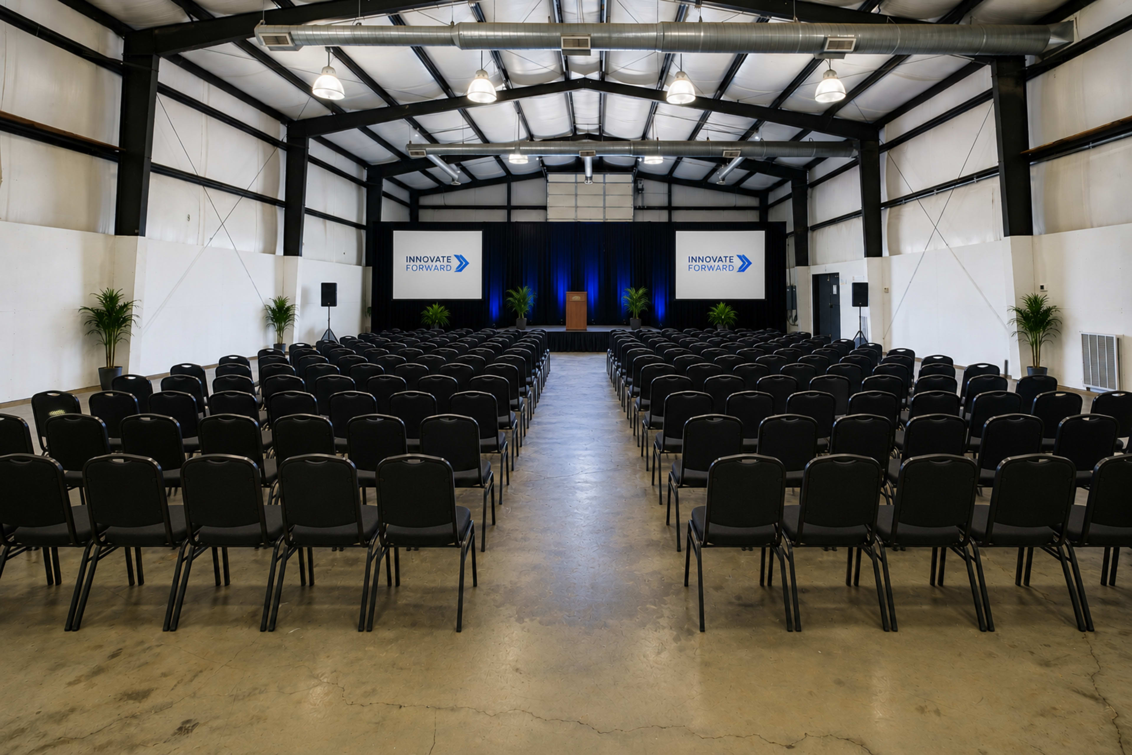 The image shows a large indoor venue set up with rows of black chairs facing a podium on a stage, flanked by two screens displaying the words "INNOVATE" and "EXPLORE."