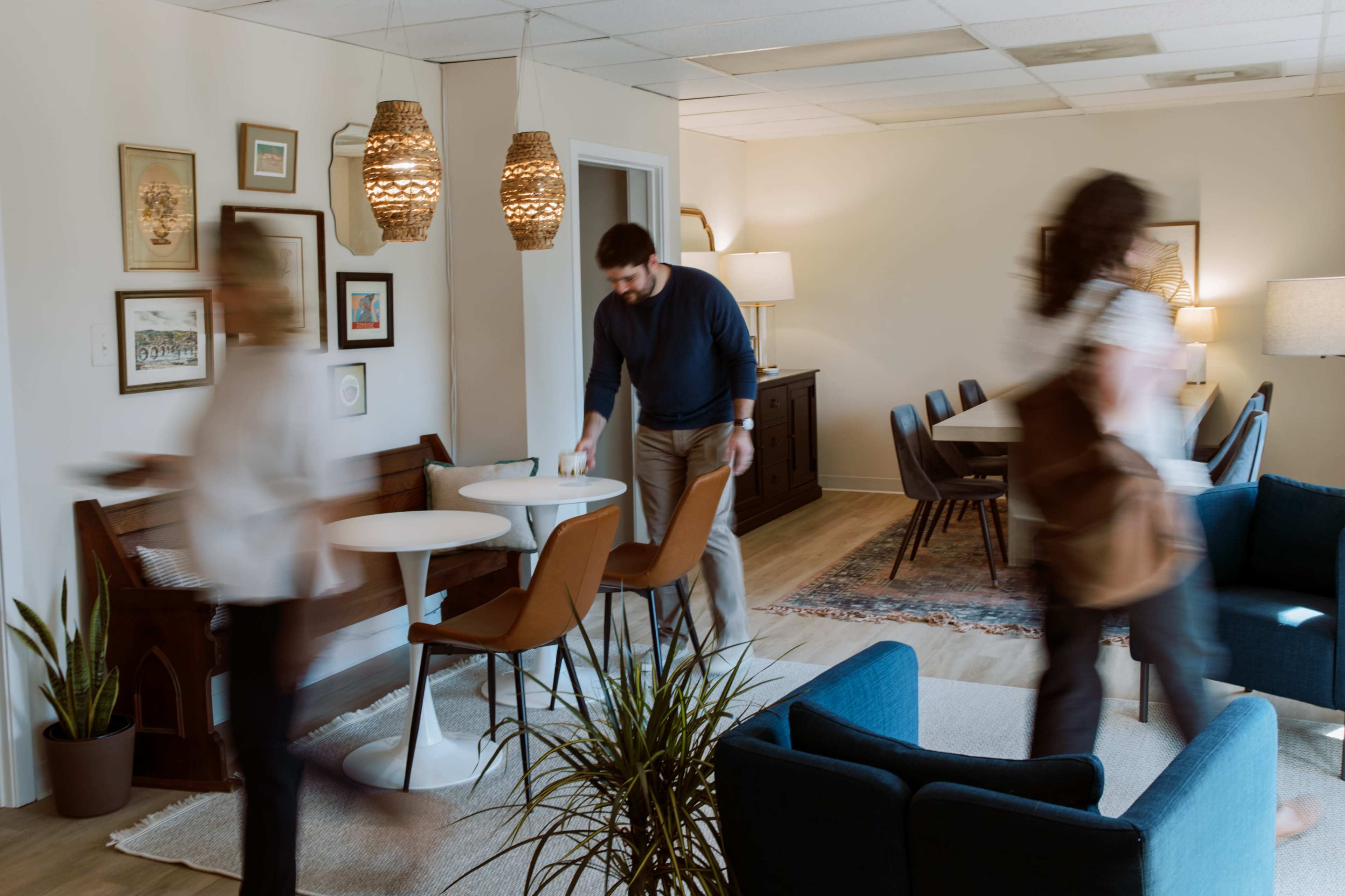 A modern office space features a dining area with a large table, casual seating, and decorative plants, as people move through the scene.
