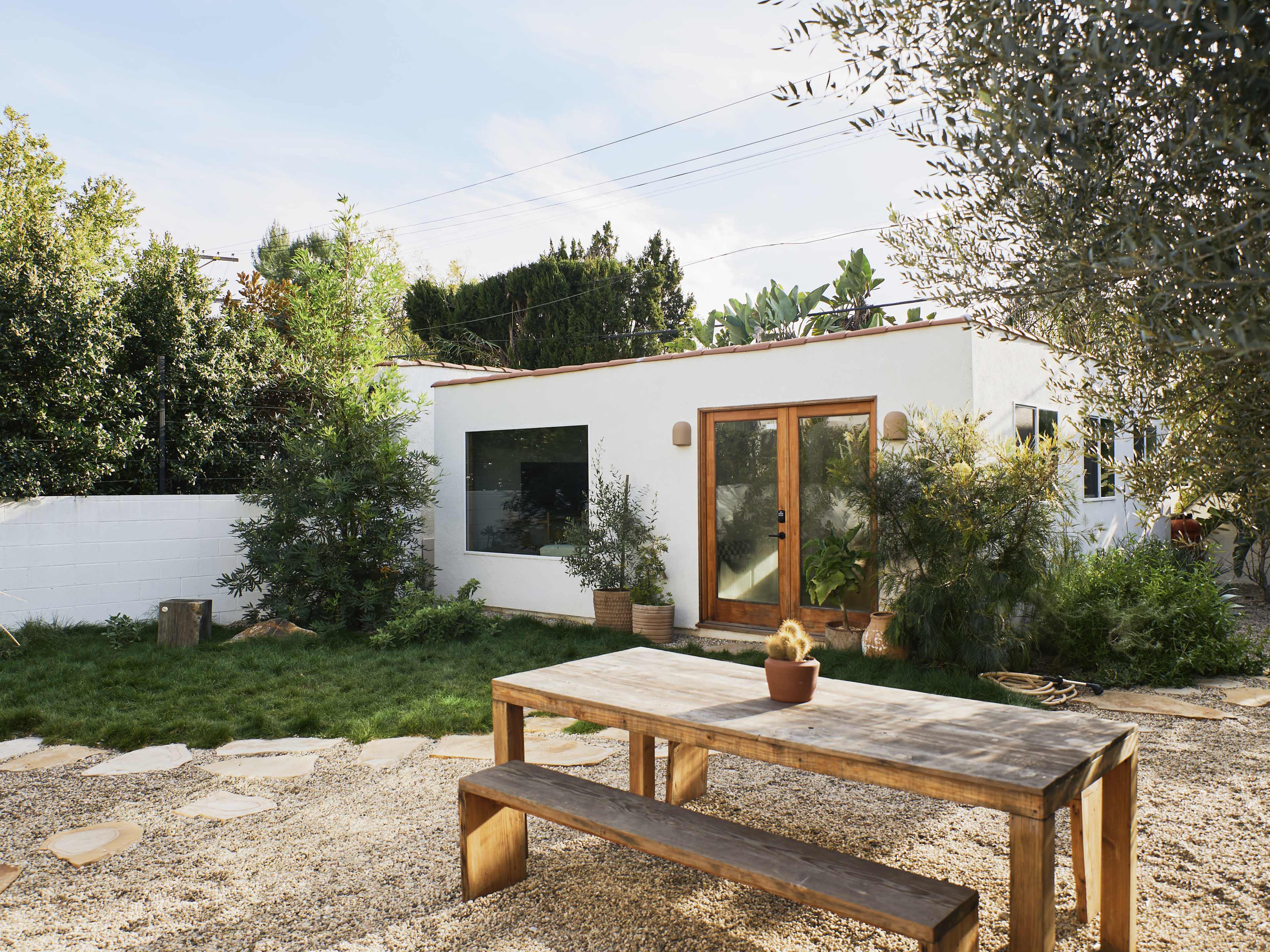 A modern, white house with large windows is surrounded by greenery and a gravel yard featuring a wooden picnic table.