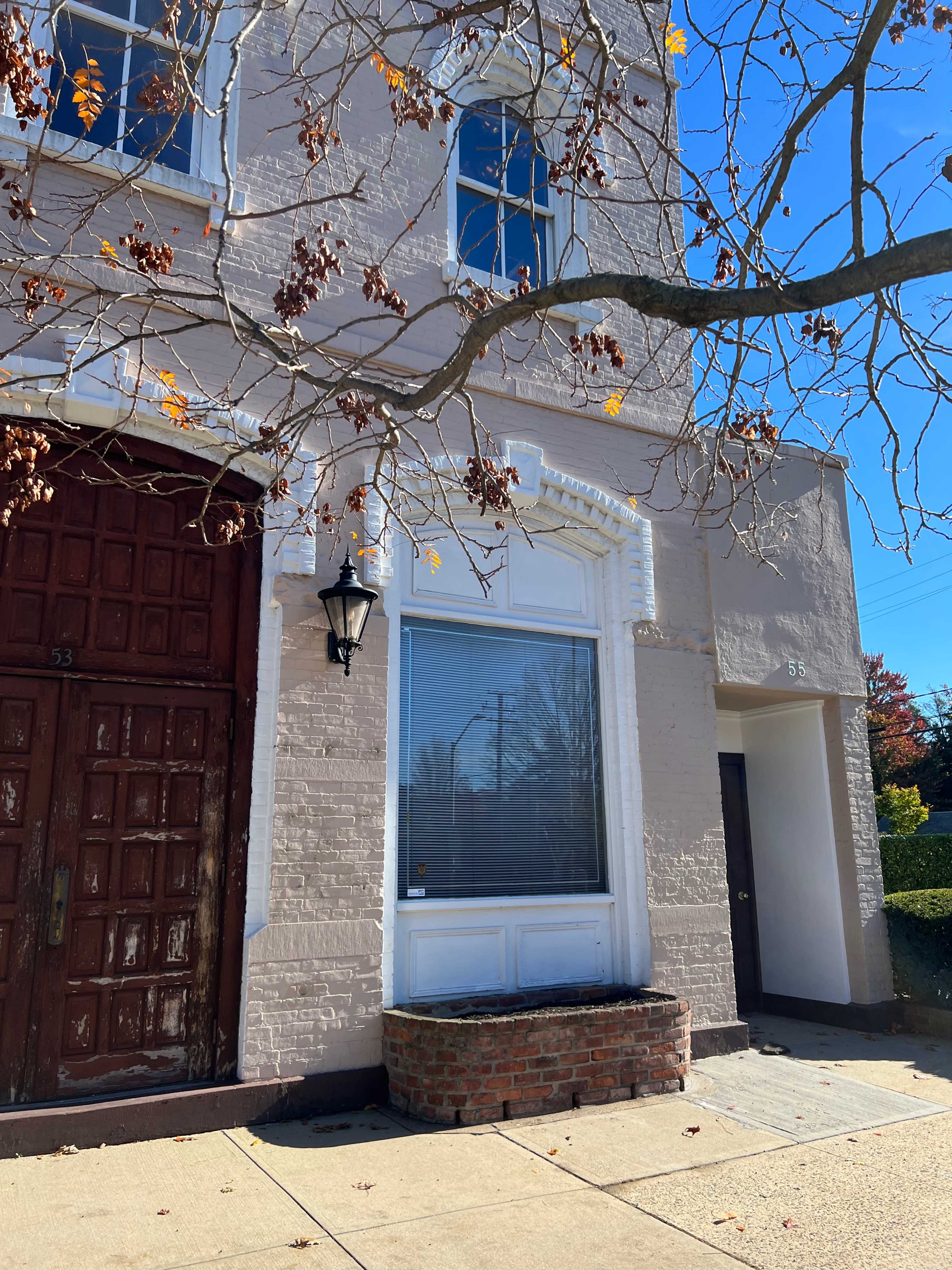 A brick building with a large window framed by a white facade and a wooden door, partially obscured by a bare tree branch.