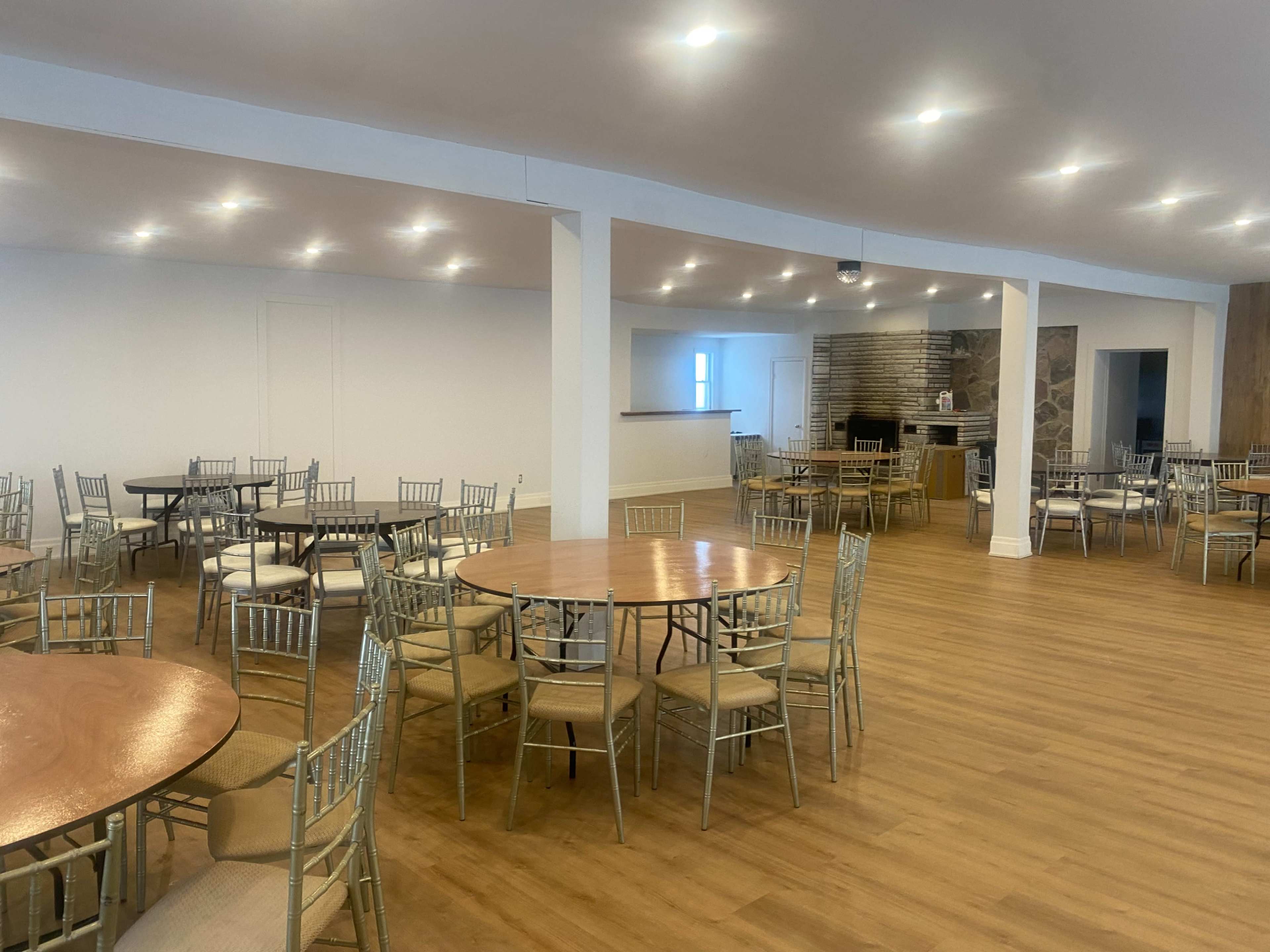 The image shows a spacious event hall with several round tables and chairs arranged on wooden flooring, featuring a stone fireplace in the background.