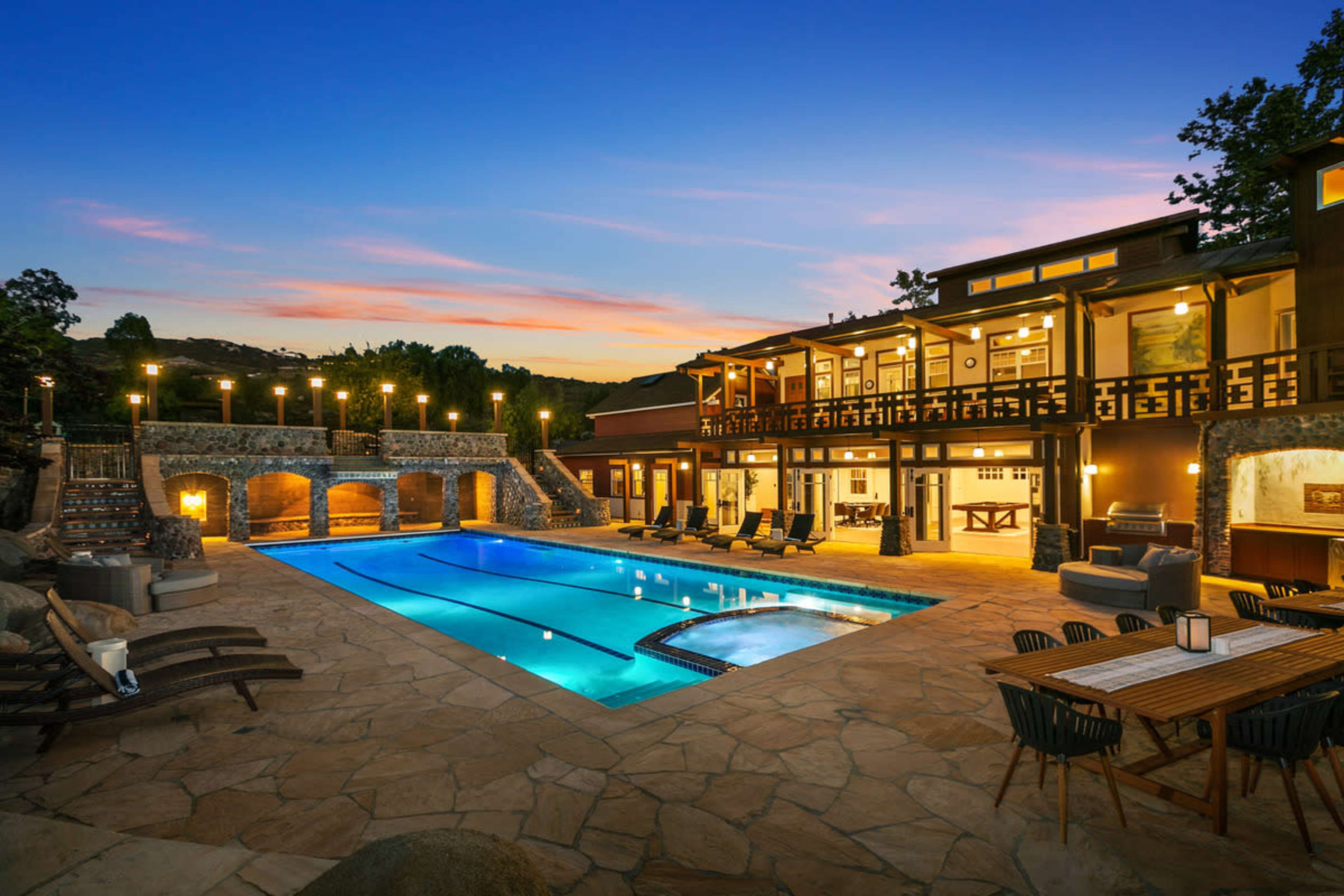 A modern villa with a large swimming pool surrounded by stone landscaping, outdoor seating, and a sunset sky.