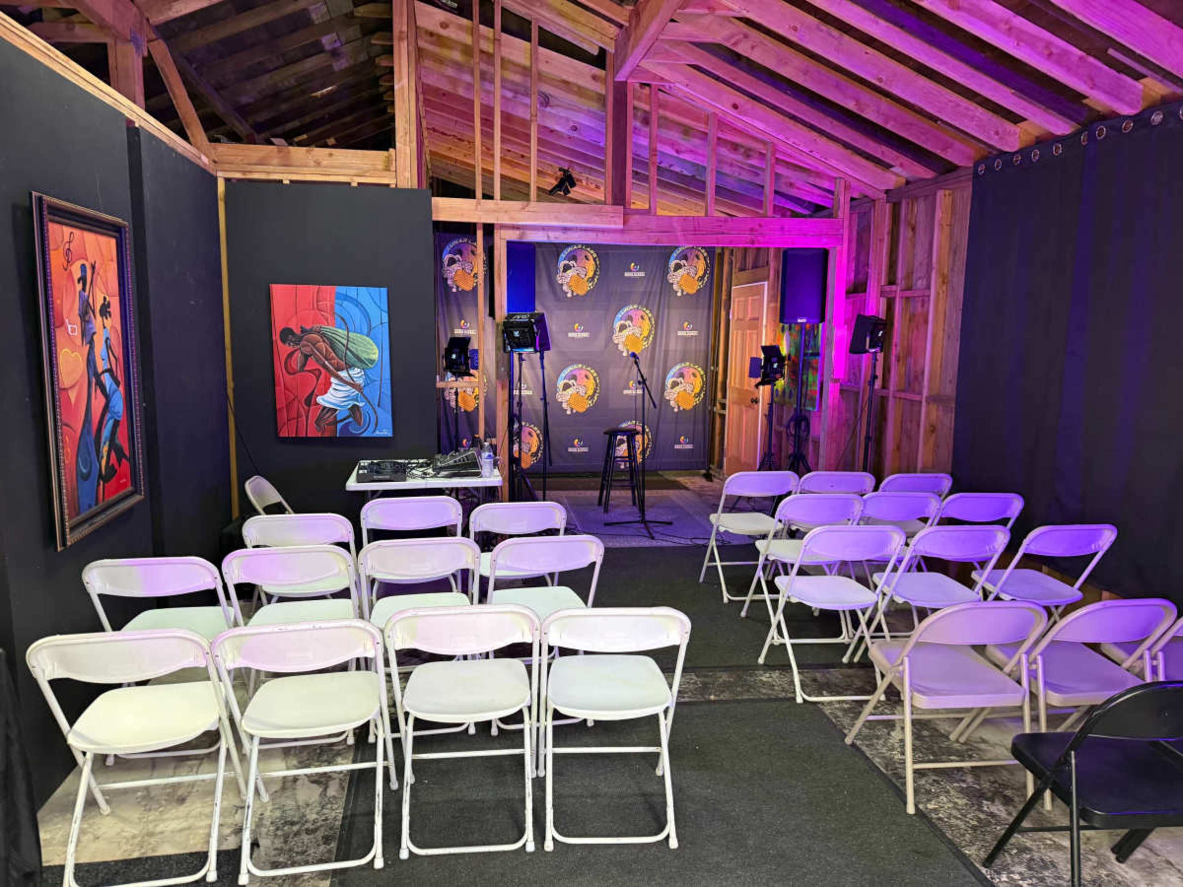 The image shows a small performance space with white chairs arranged in rows facing a microphone on a stool, flanked by speakers and a decorative backdrop.