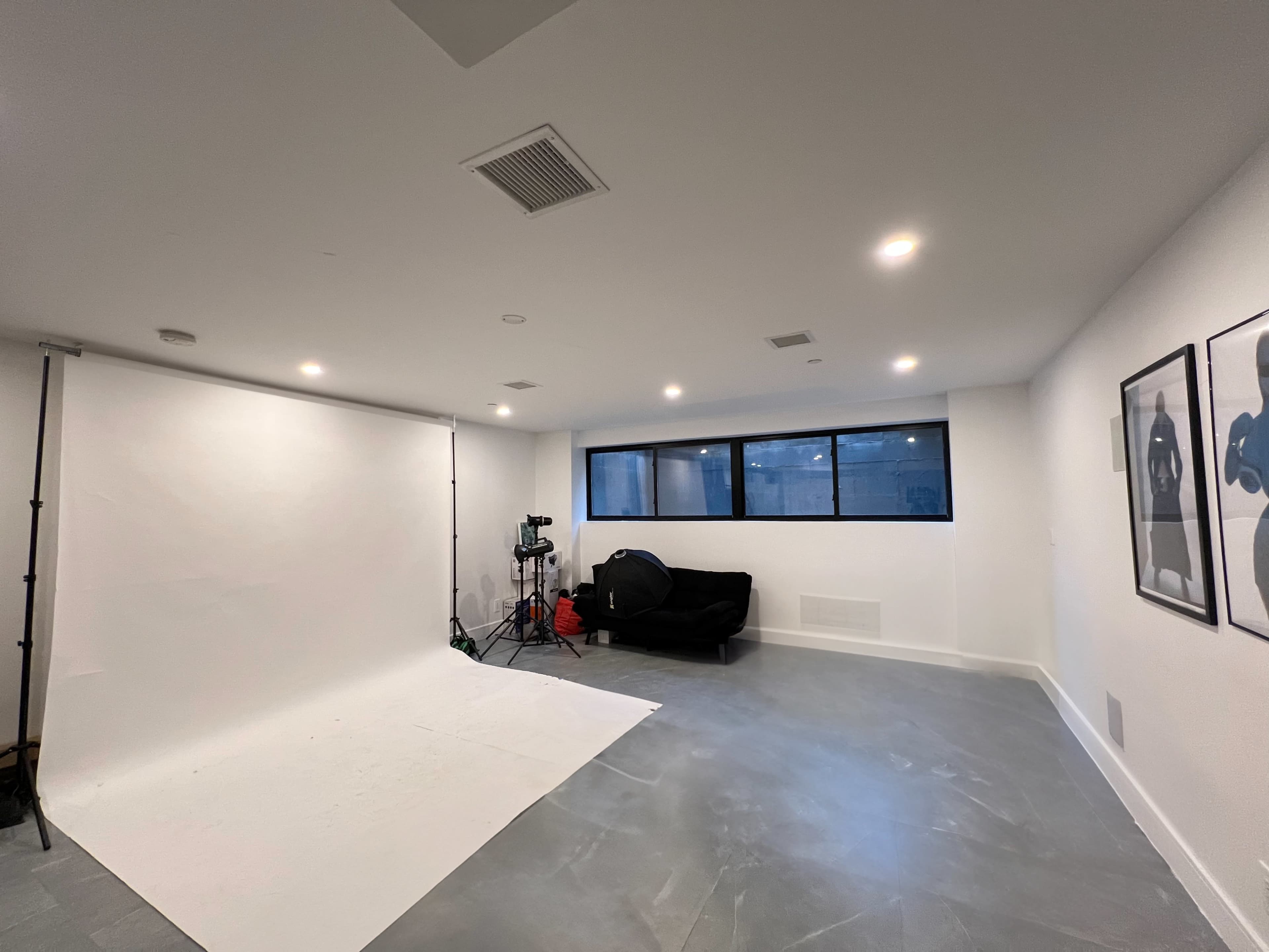The image shows a spacious, well-lit room with a white backdrop, a black sofa, and large windows.