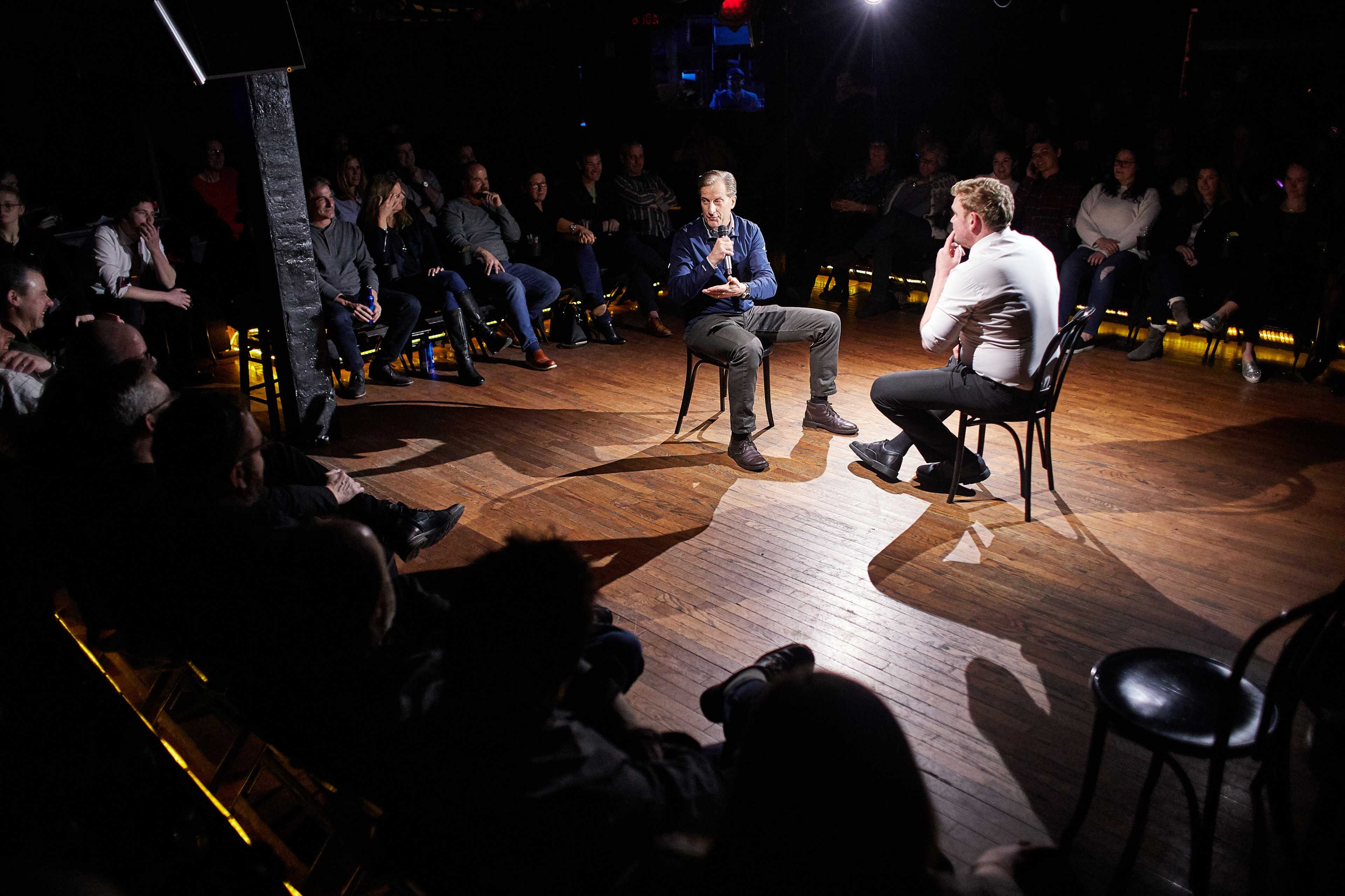 Two individuals are seated on chairs facing each other on a dimly lit stage in front of an audience.