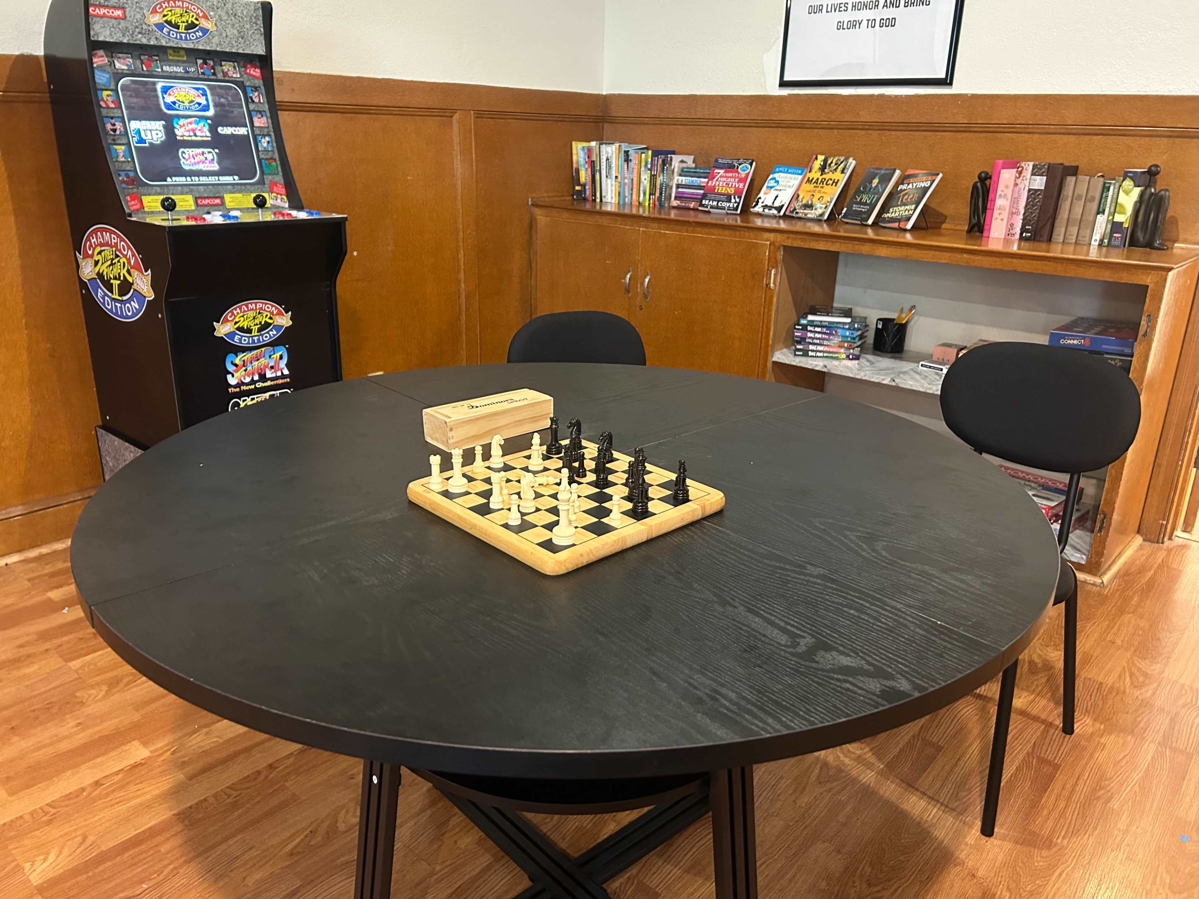 The image shows a round black table with a wooden chess set in the center, surrounded by two black chairs, and an arcade machine along with shelves of books in the background.