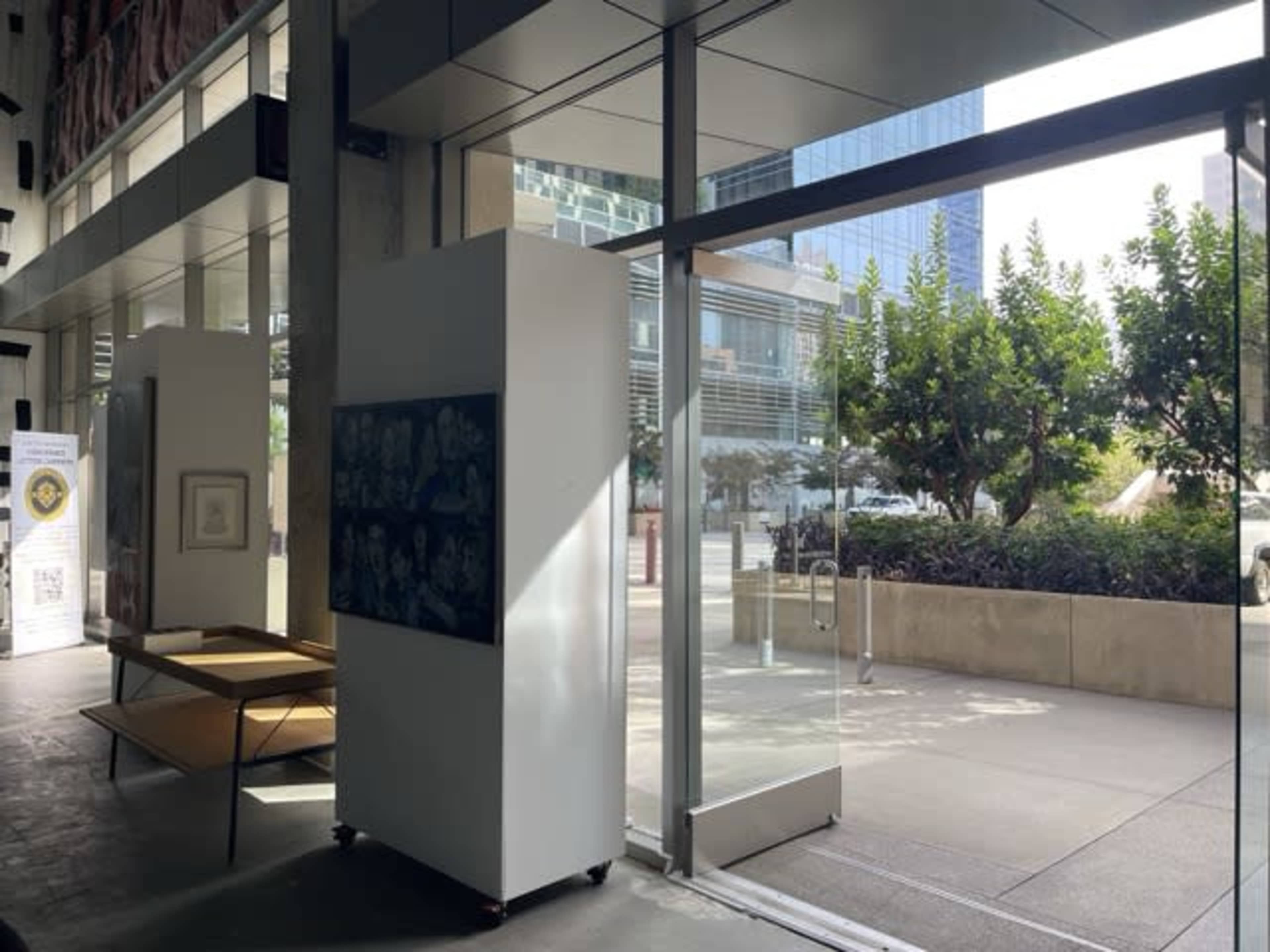 The image shows a modern interior space with large glass doors leading to a landscaped area outside, featuring a display of art on the wall and a table nearby.