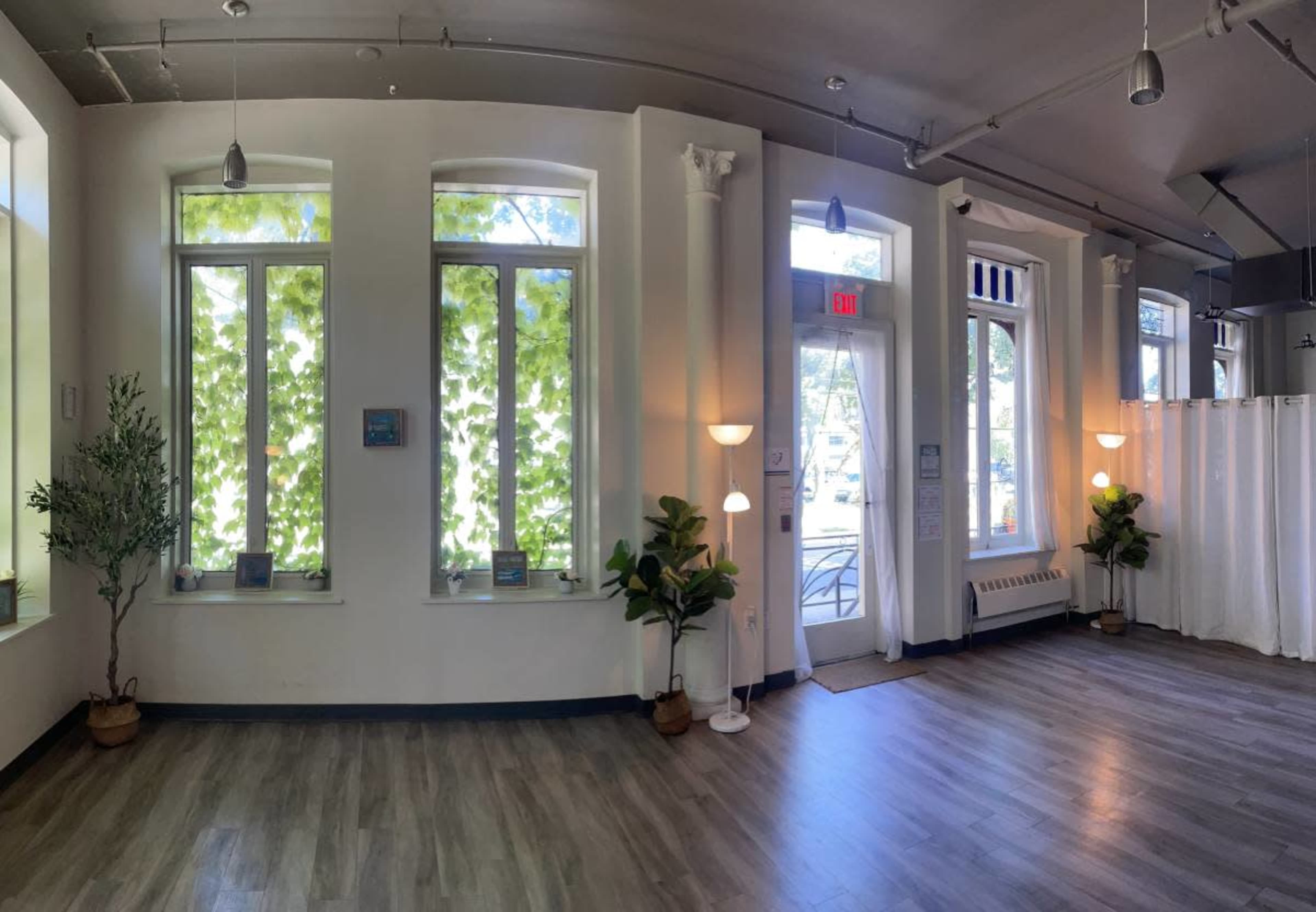 A spacious interior with large windows letting in natural light, potted plants in each corner, and an entrance door leading outside.