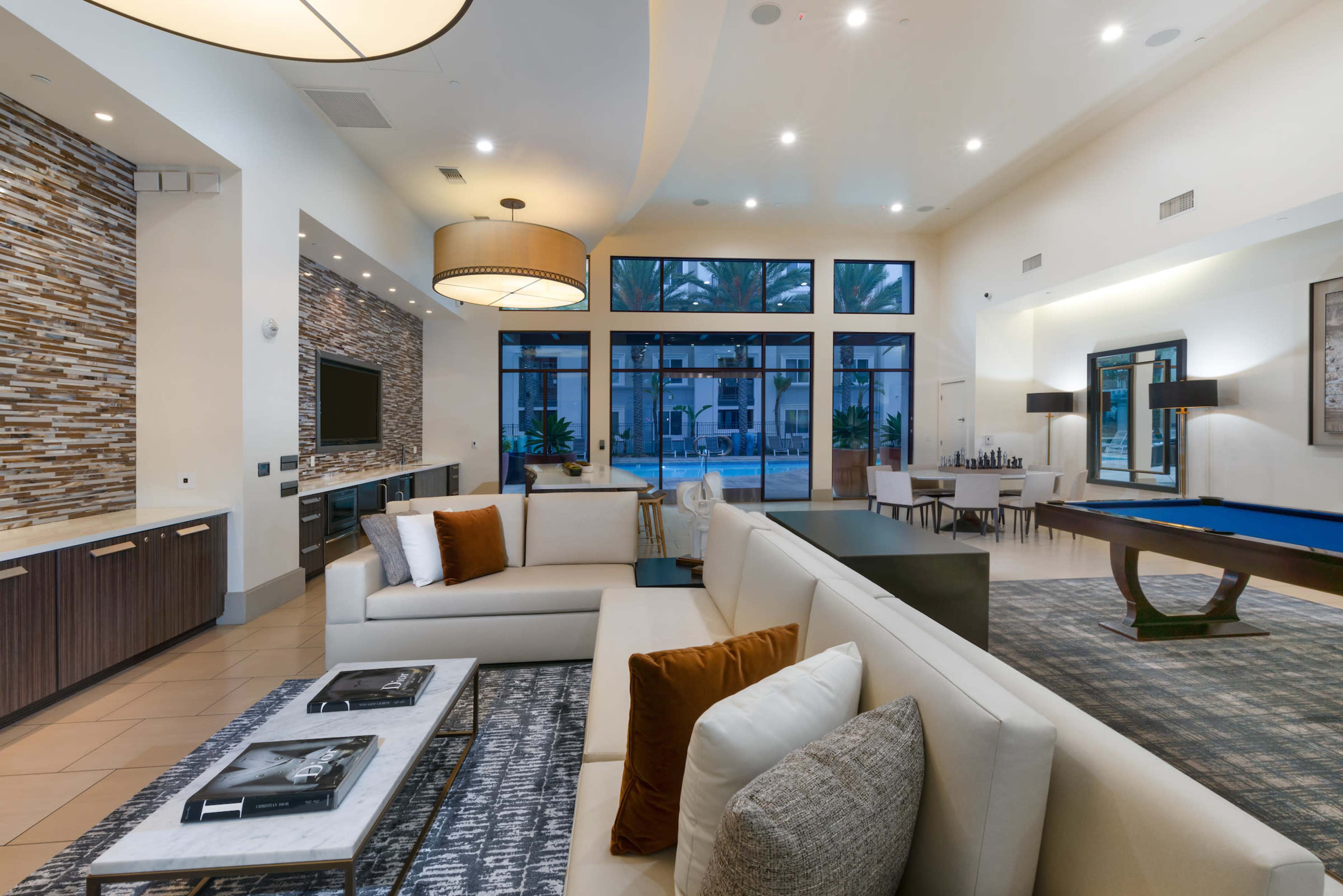 A modern living space featuring a sectional sofa, a coffee table, a pool table, and large windows overlooking an outdoor area.