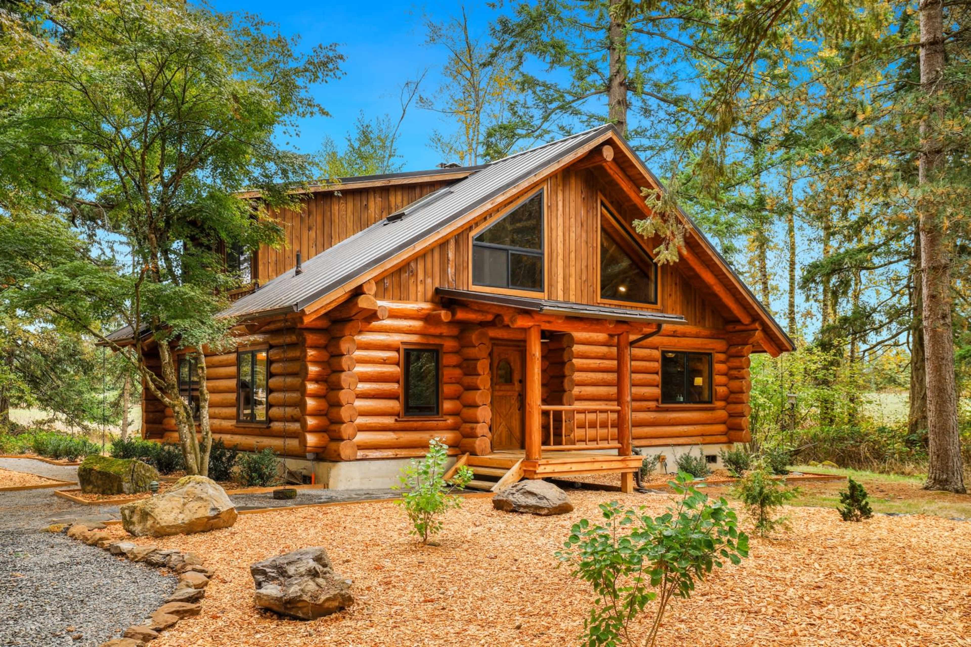 The image shows a rustic log cabin surrounded by trees and landscaped with rocks and gravel paths.