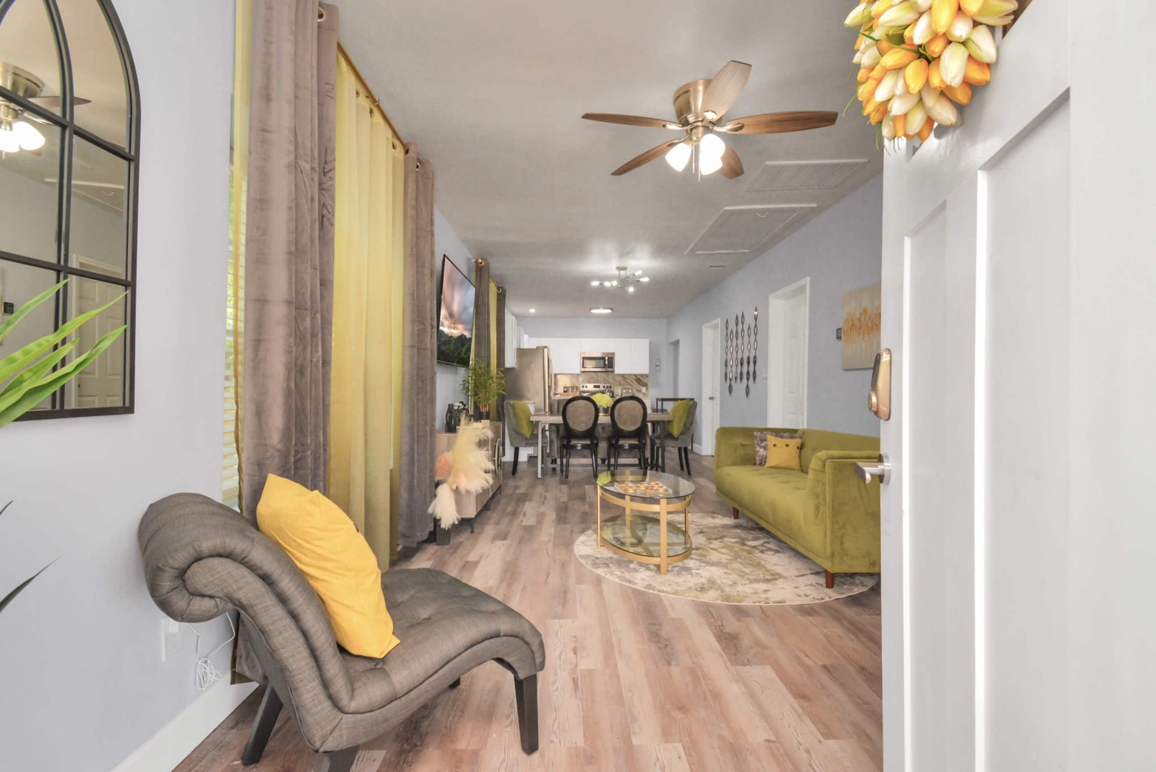 The interior of a modern living space features a green sofa, a round coffee table, and various decorative elements, leading to a dining area in the background.