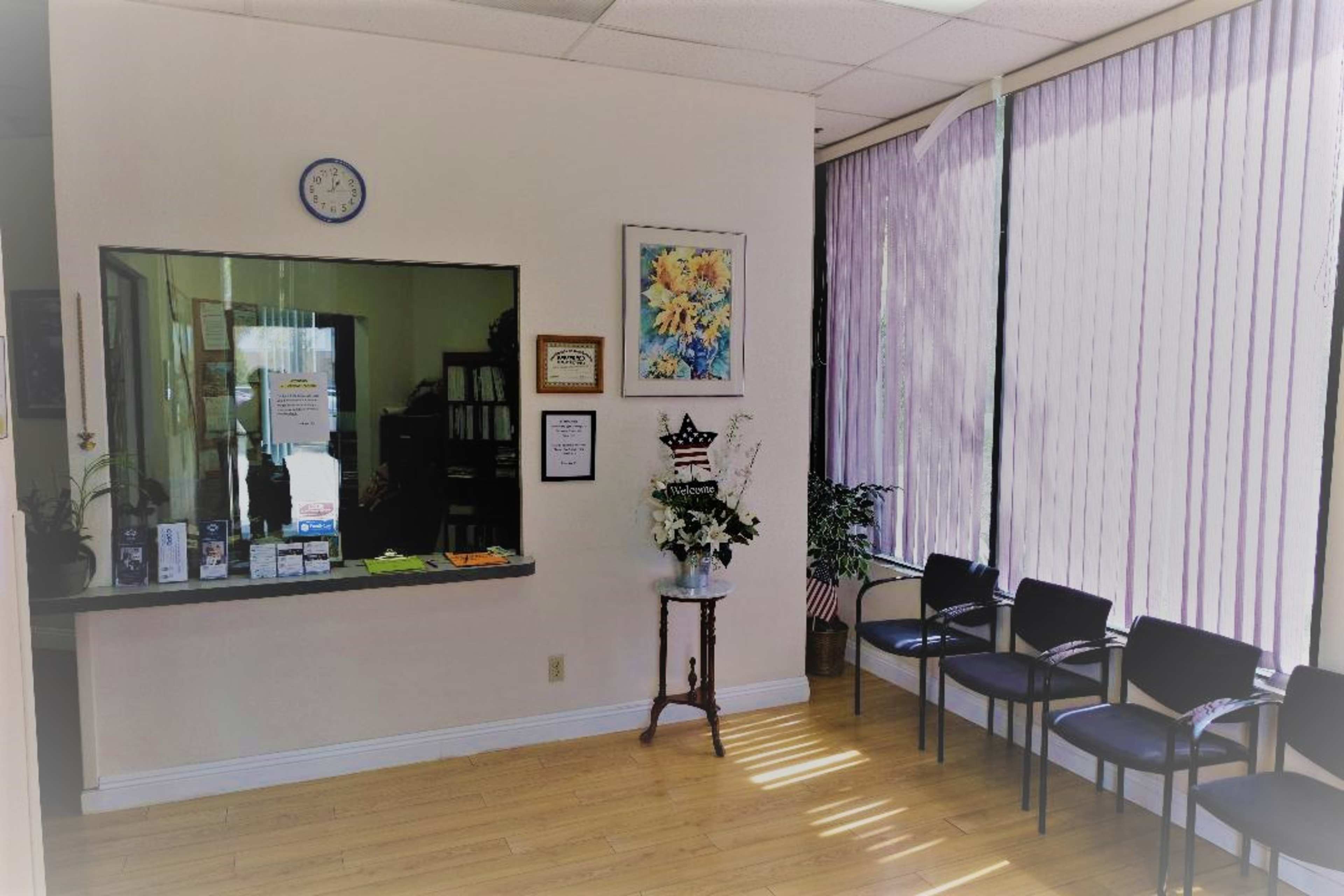 A waiting room with a counter, a floral display, and several black chairs arranged in front of large windows with purple curtains.