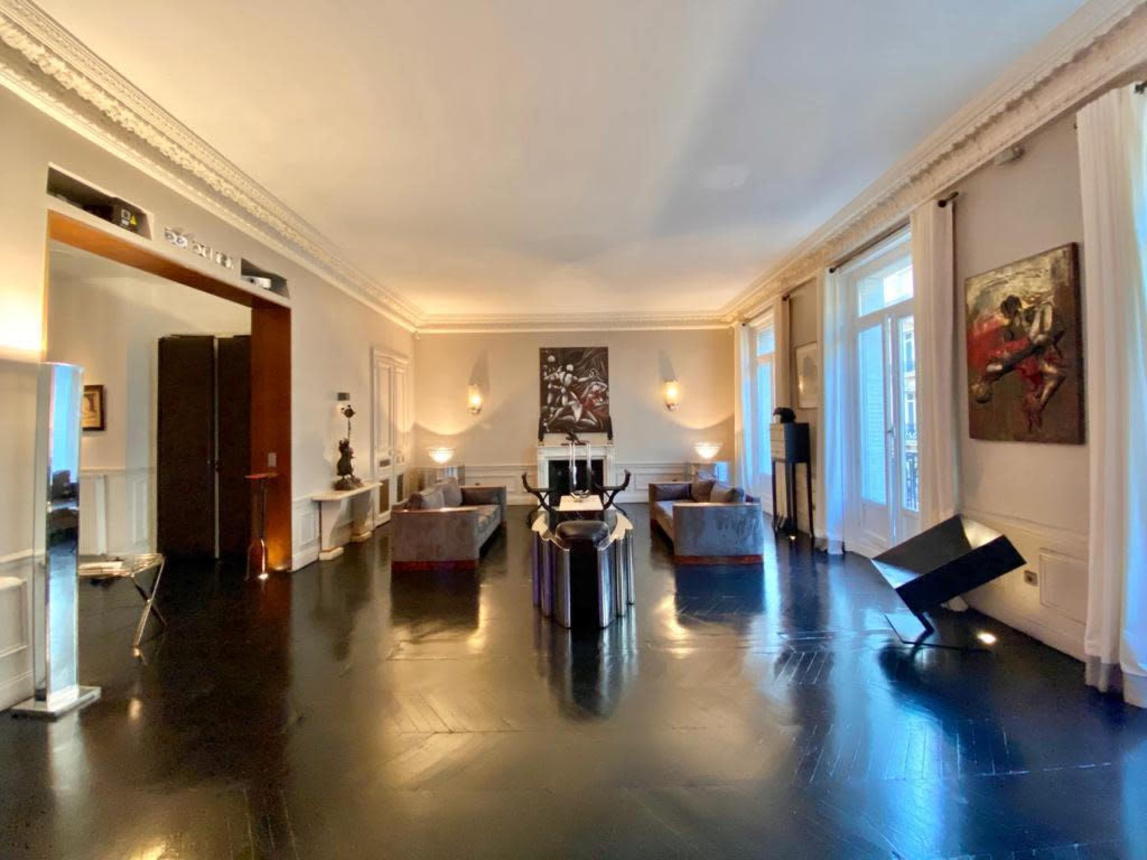 The image shows a spacious living room with dark wooden floors, large windows, and contemporary furniture arranged around a central seating area.