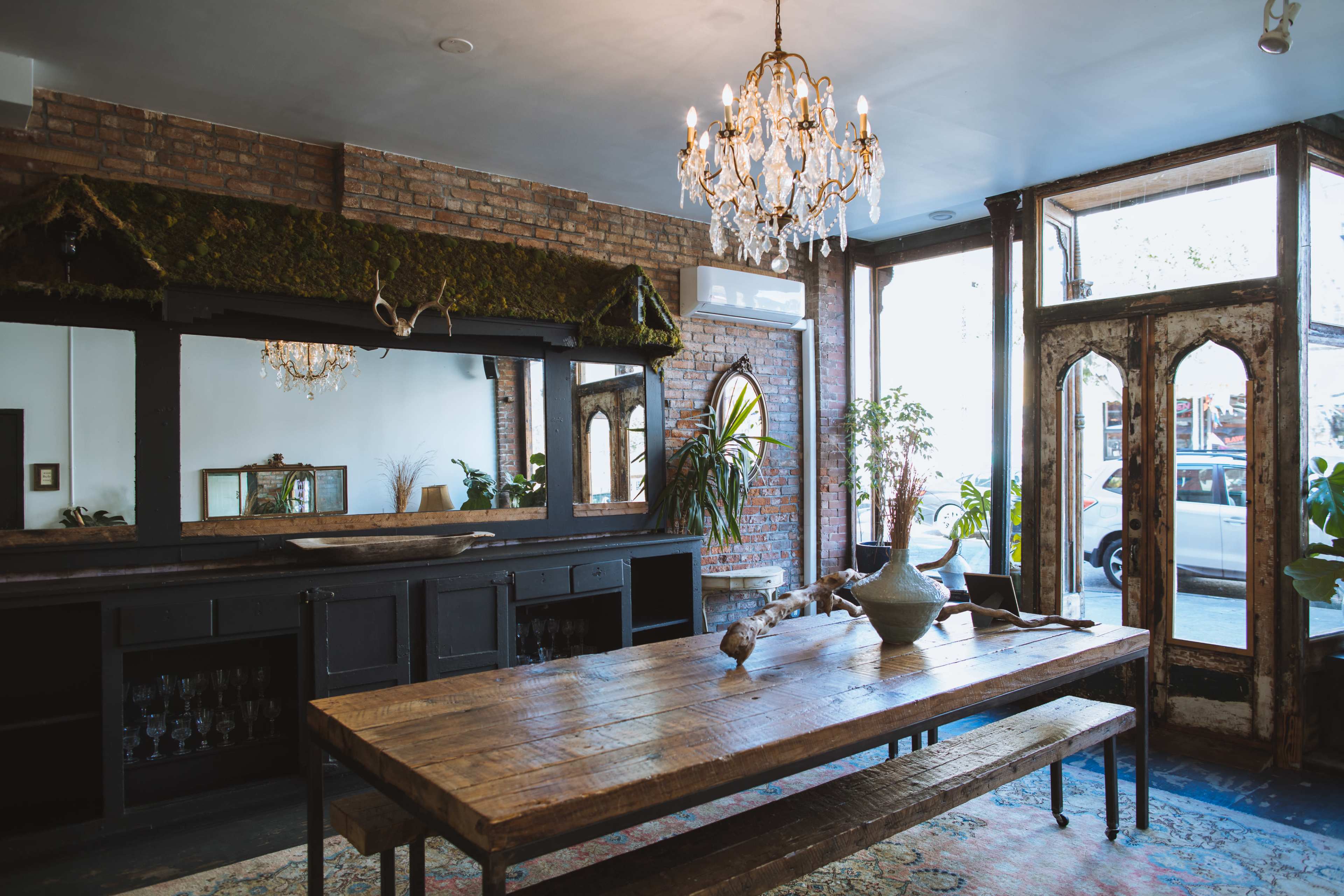 An interior space featuring a wooden dining table, a vintage chandelier, and a rustic mirror, with large windows letting in natural light and plants adding greenery to the scene.