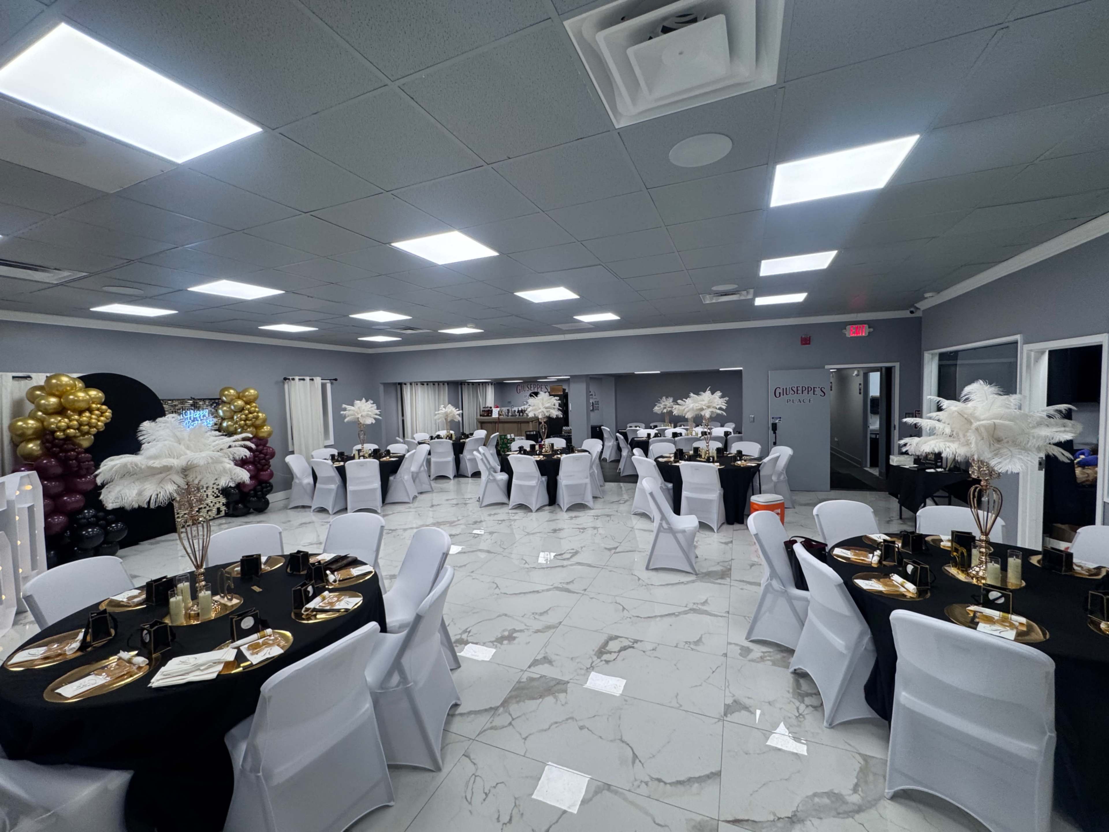 The image shows a banquet hall decorated with round tables covered in black tablecloths, white chairs, and centerpieces featuring white feathers.